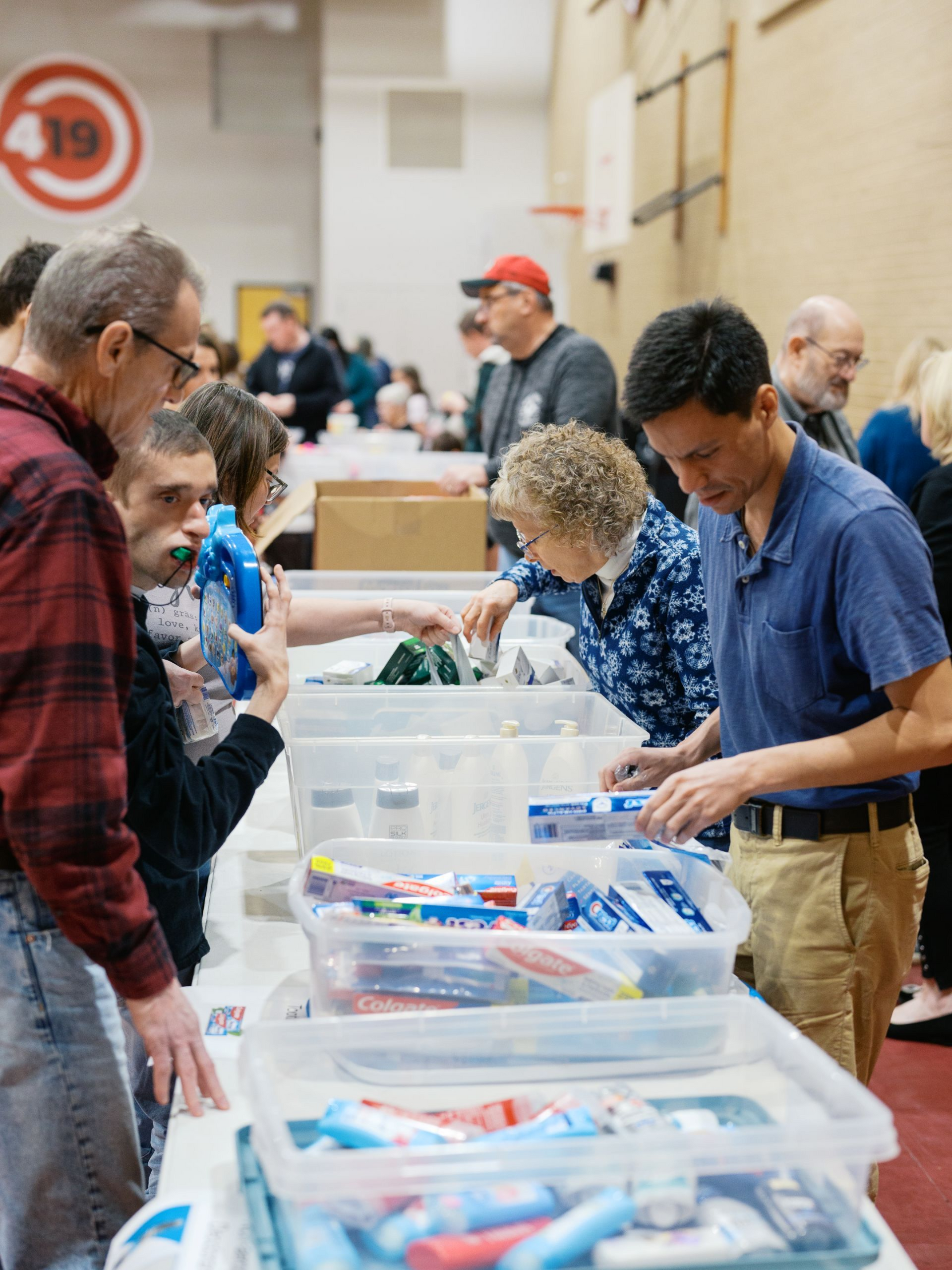People browsing bins of items at an indoor community event, with volunteers assisting nearby.
