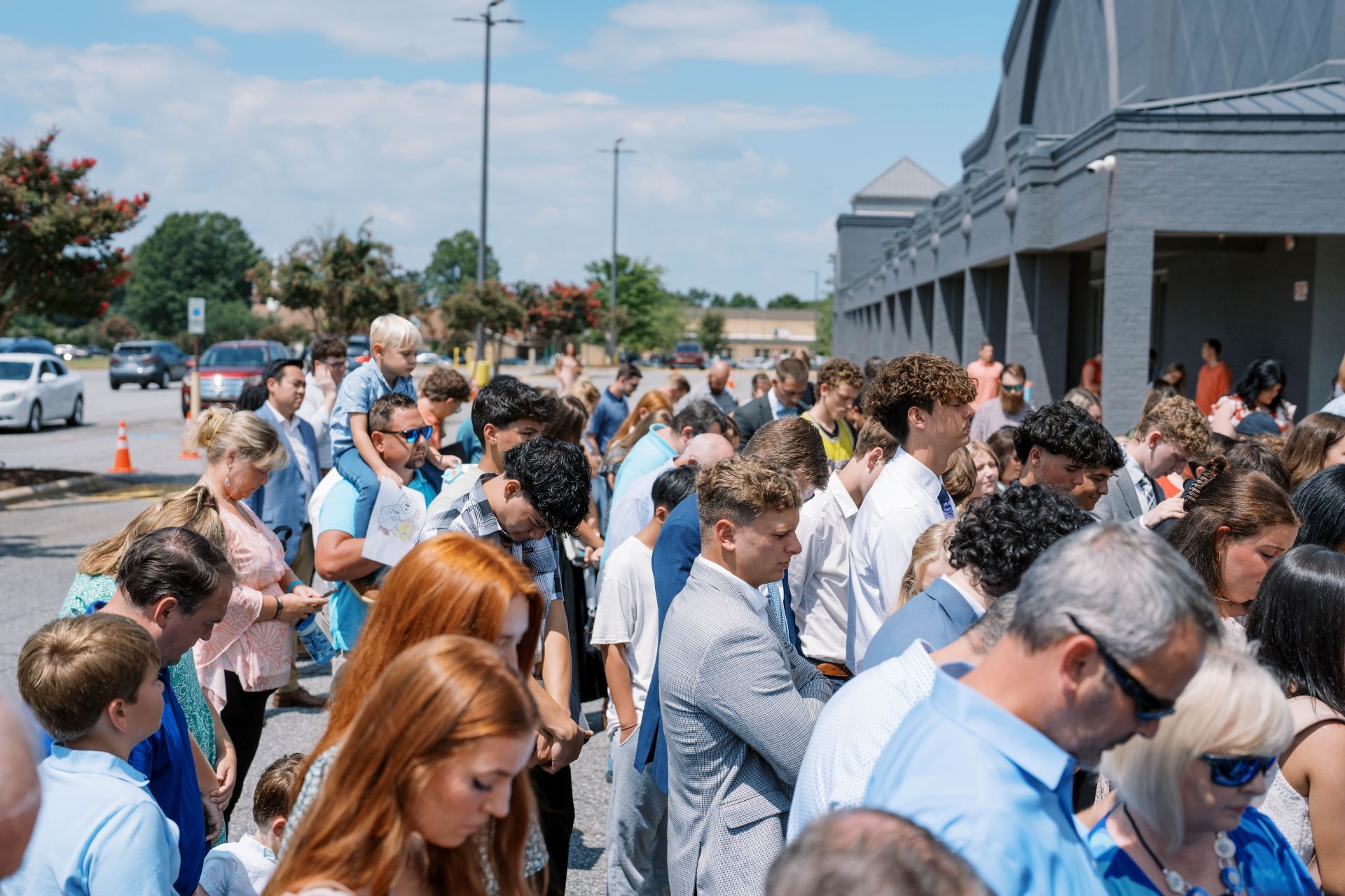 Crowd gathered outside a building on a sunny day, many people looking downward and waiting.