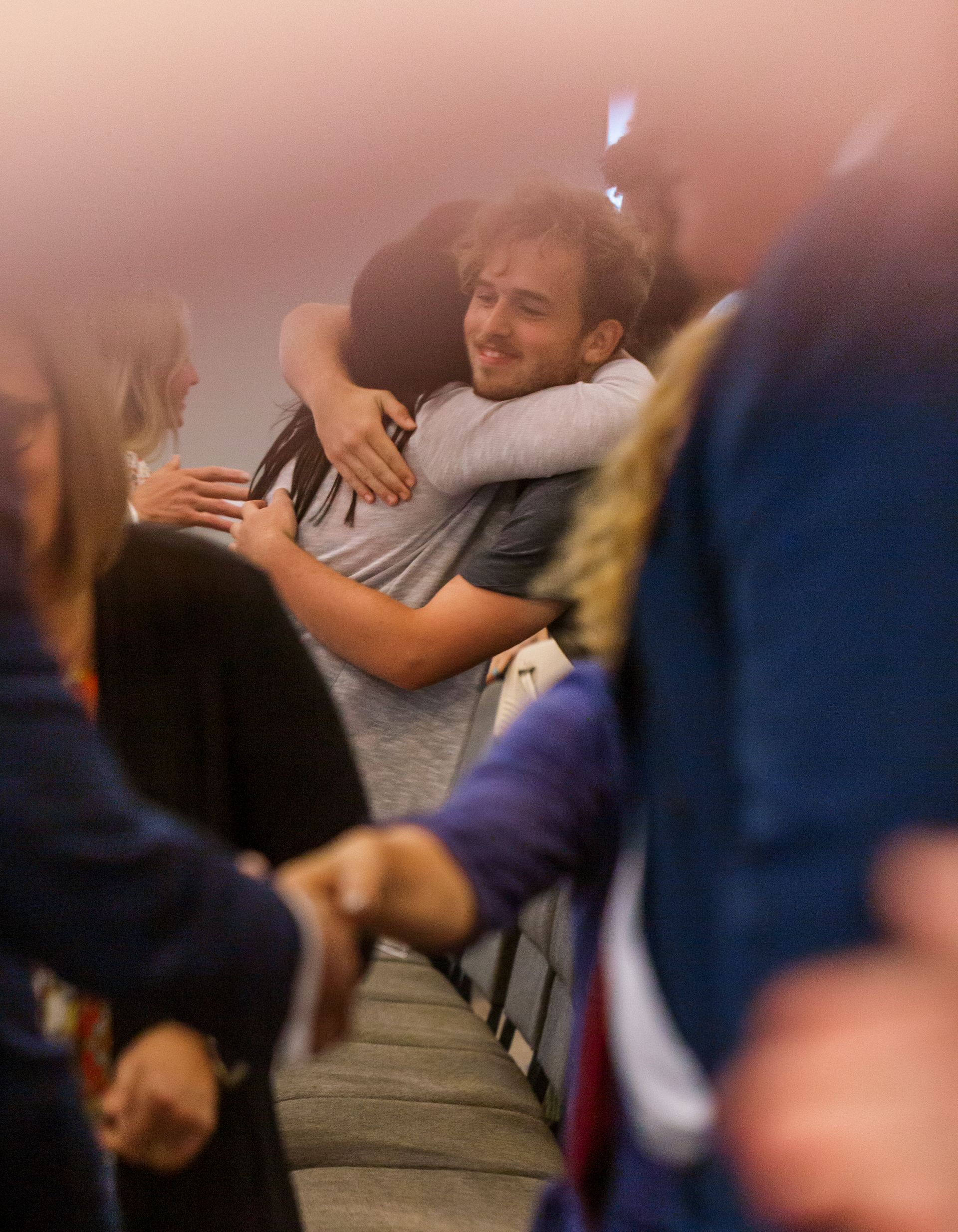 Two people hugging in a crowded indoor hallway, one smiling while others blur past in the foreground.