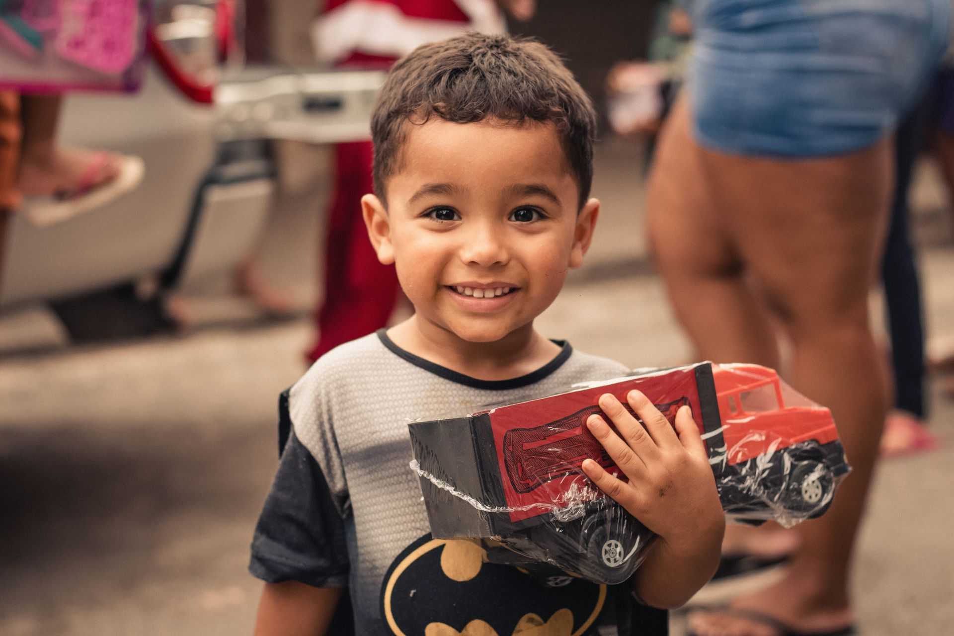 Smiling child holding a red toy truck indoors, with people blurred in the background