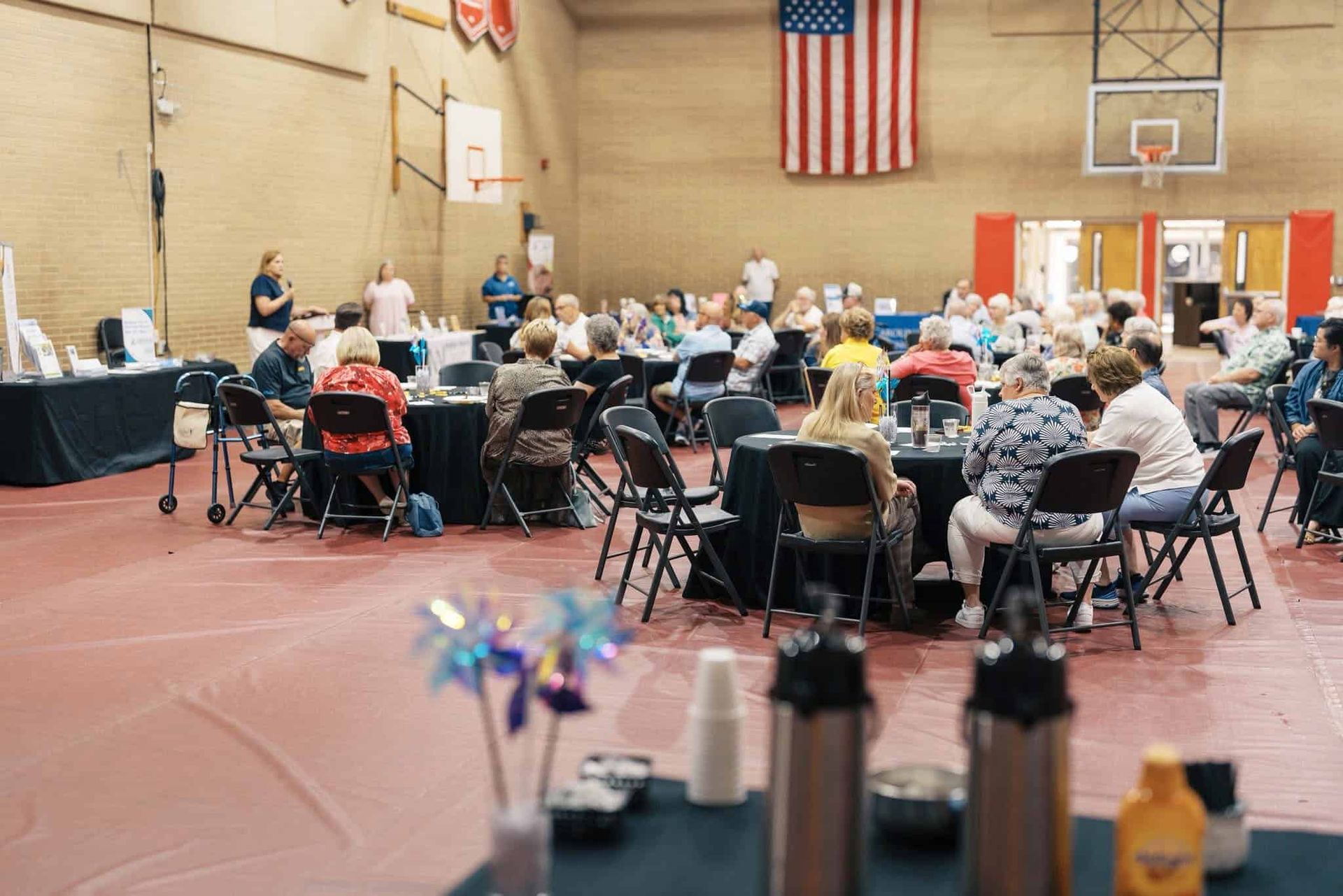 Community banquet in a gymnasium with people seated at round tables beneath a large American flag.