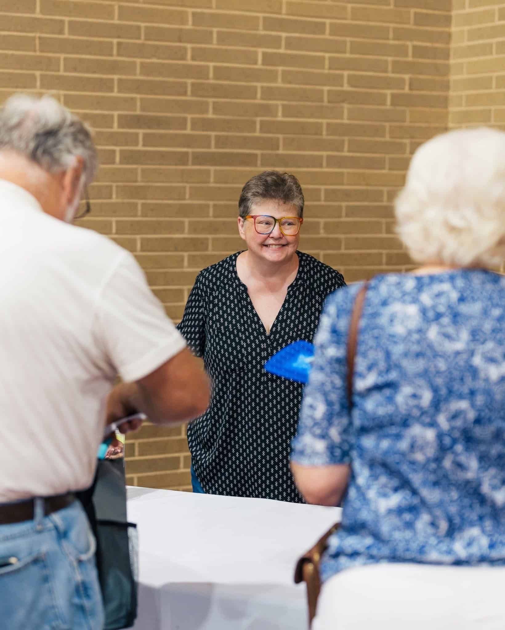 Woman in black dress with blue folder speaks to a small group indoors near a brick wall.