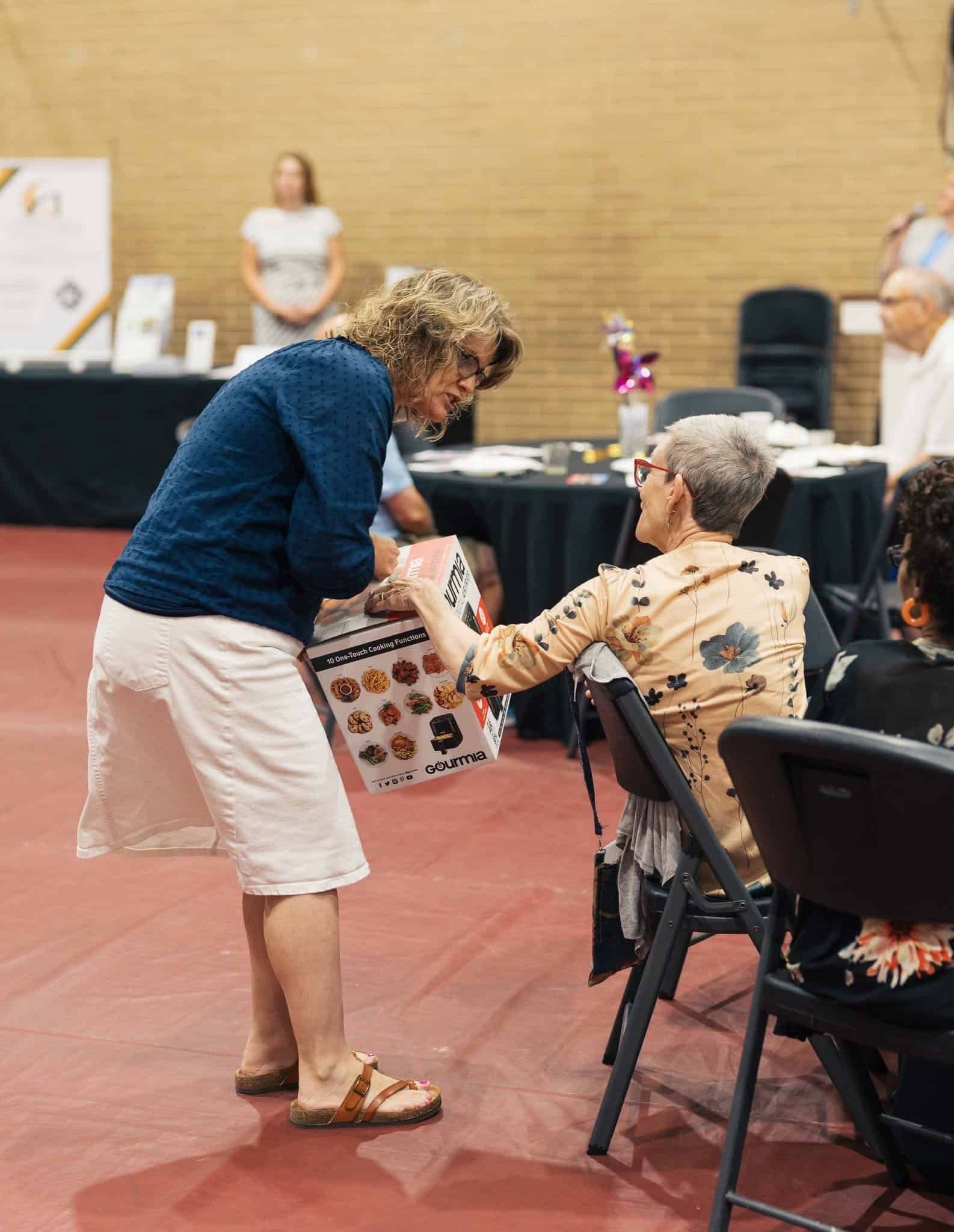 Woman handing a booklet to an elderly seated person at an indoor community event