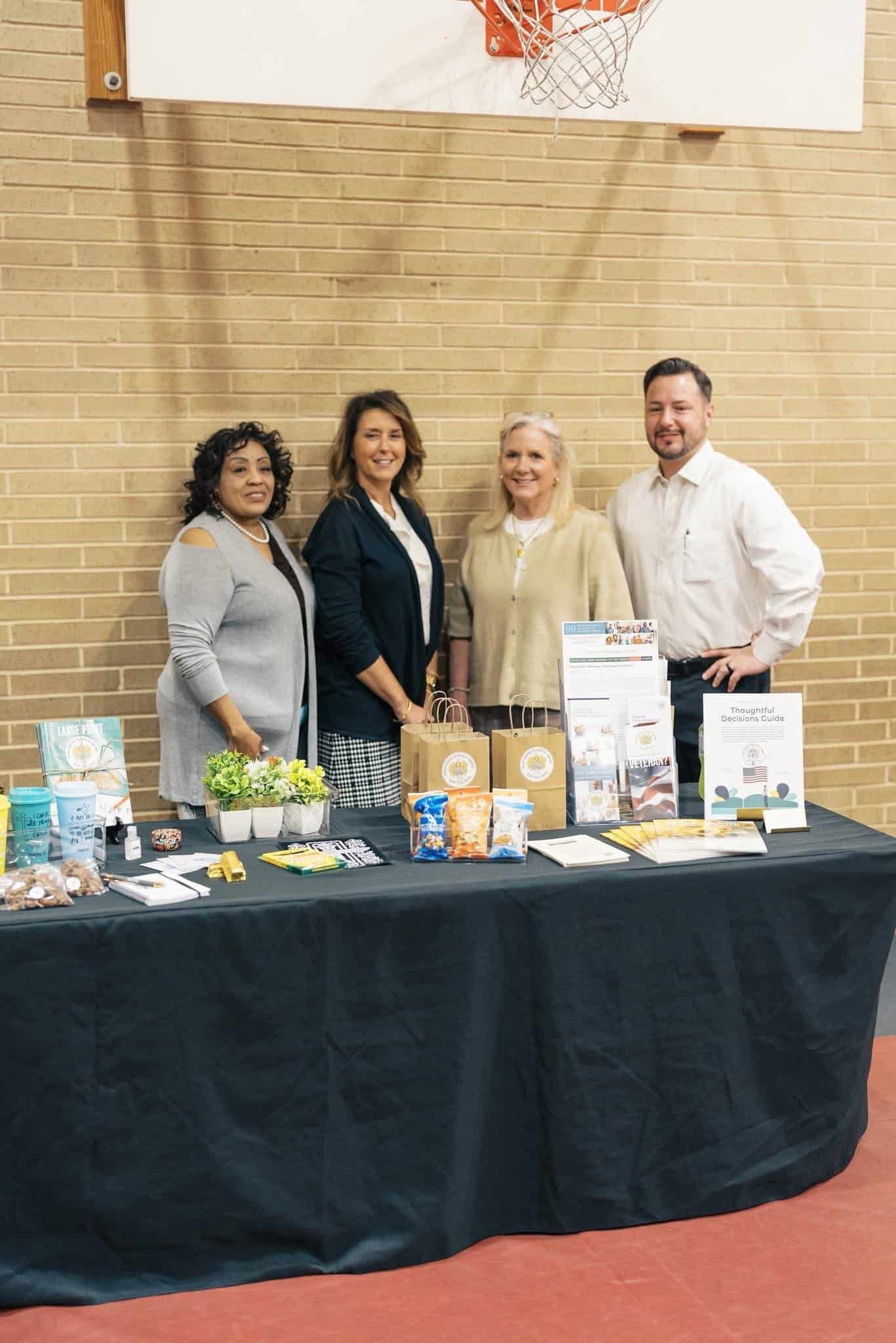 Four people standing behind a display table with flyers and products in a gymnasium or event hall