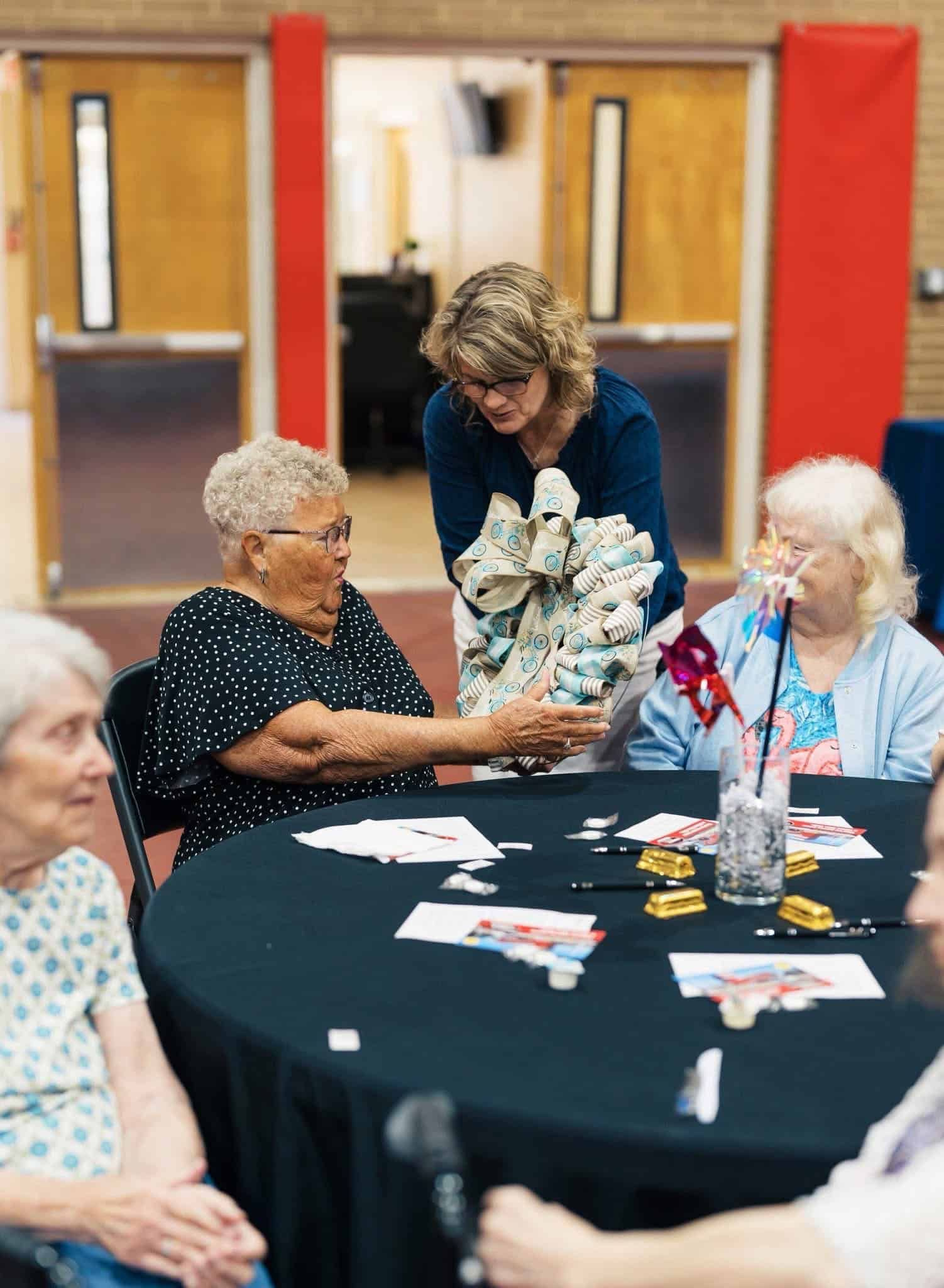 Several people at a table with papers and flowers, chatting in a community room