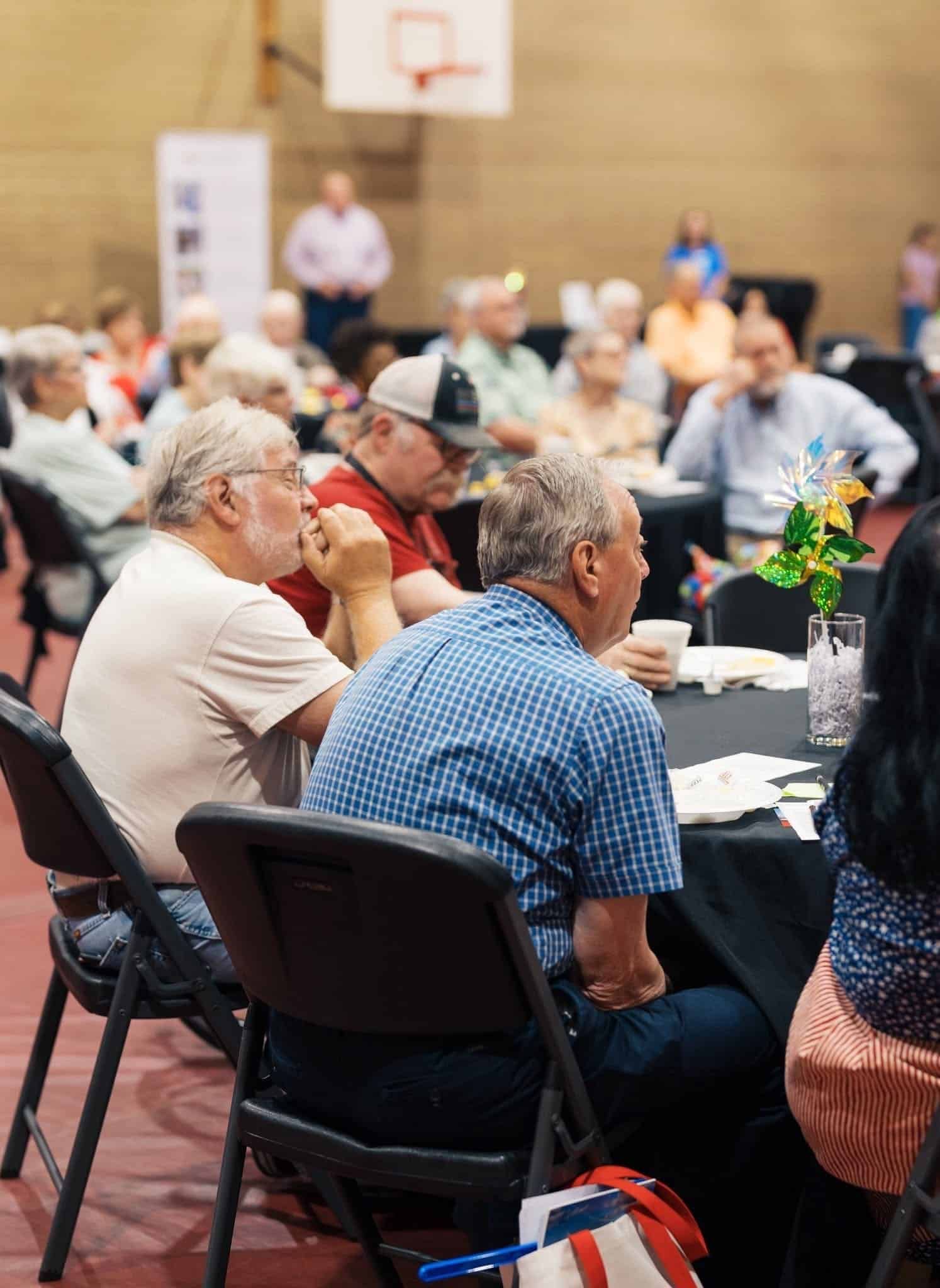 People seated at round tables in a gymnasium, listening at a community event