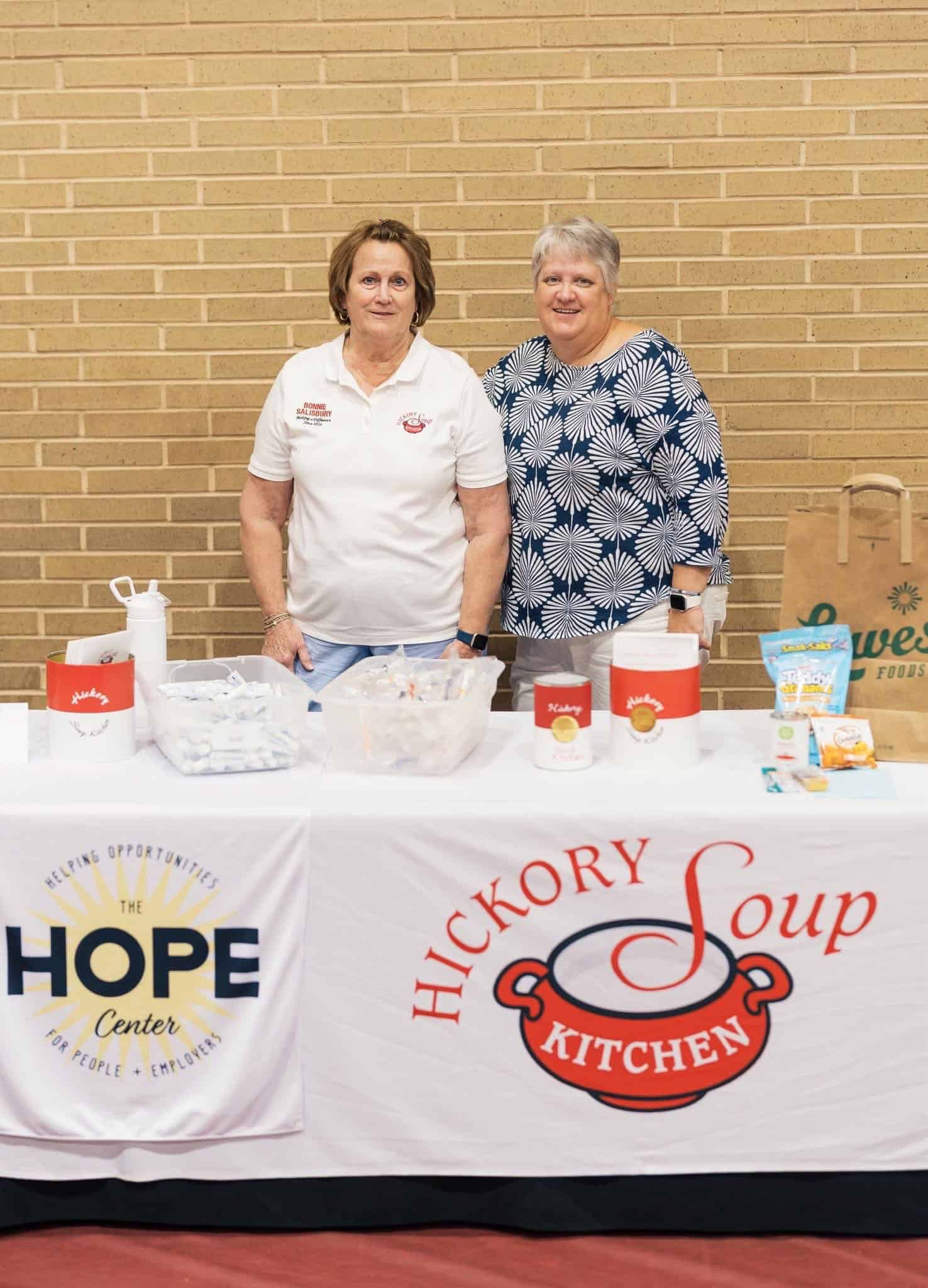 Two women behind a food table with HOPE and Hickory Soup Kitchen banners