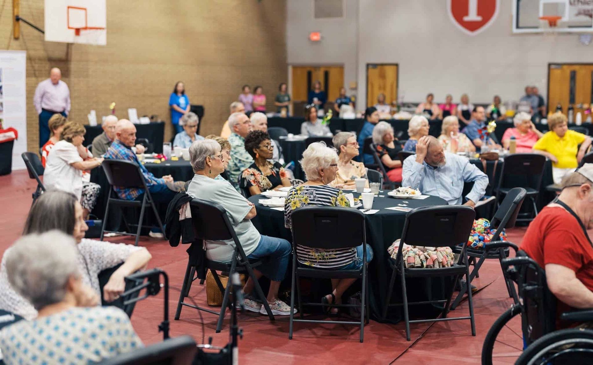 Audience seated at round tables in a gymnasium, watching an event near a basketball hoop.