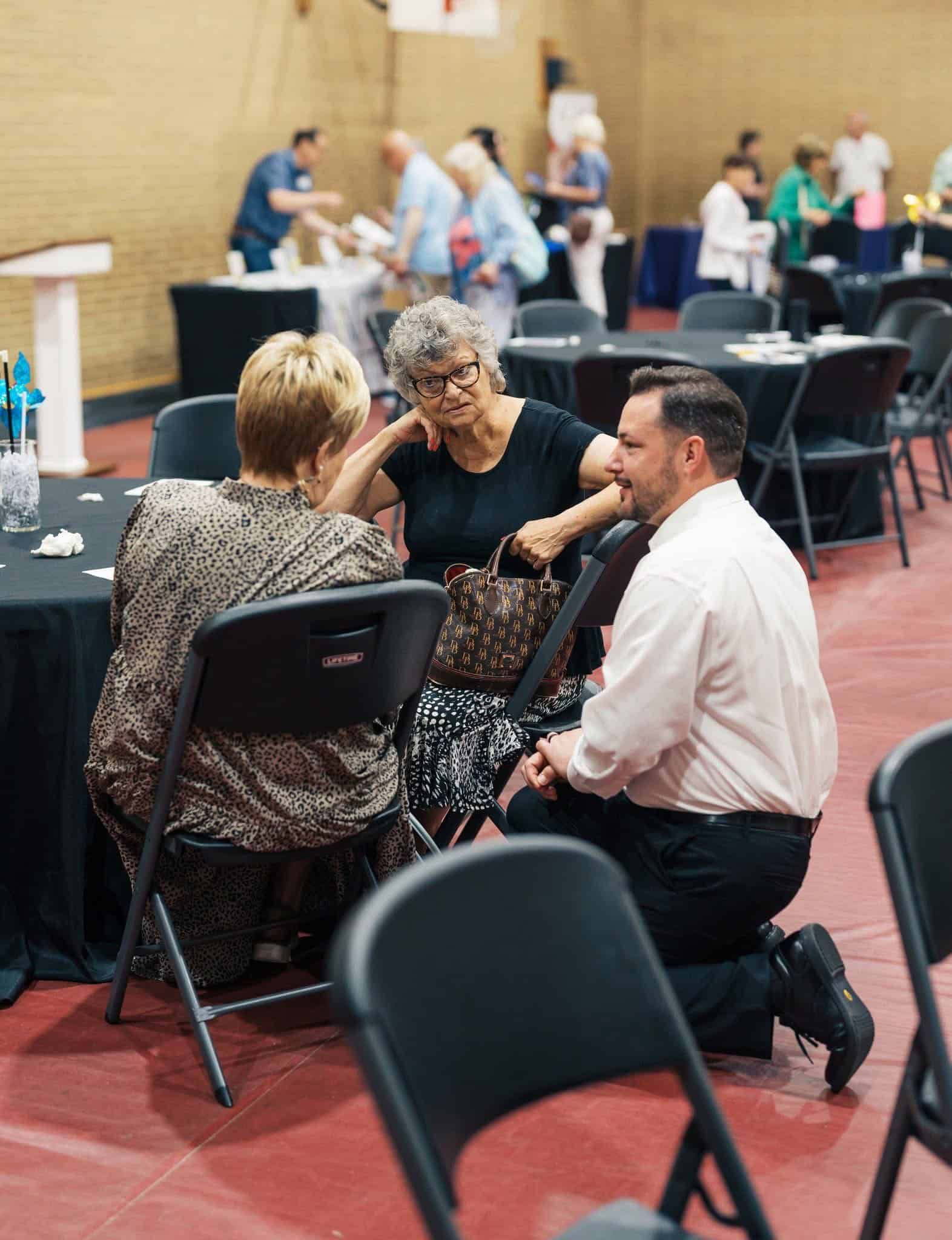 People chatting at round tables in a banquet hall, with a man kneeling beside two seated guests.