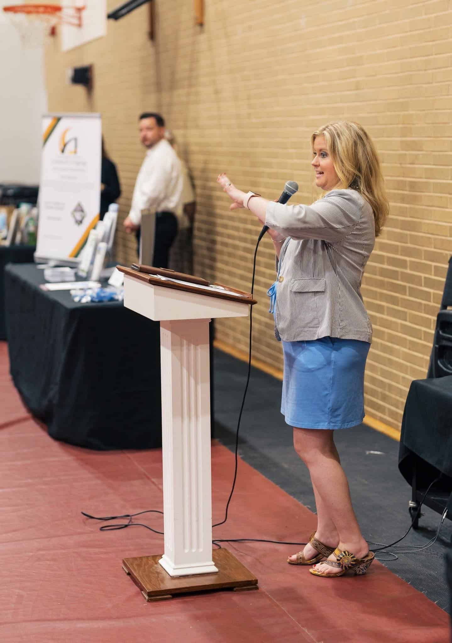 Woman speaking at a podium in a gymnasium, gesturing toward the audience.