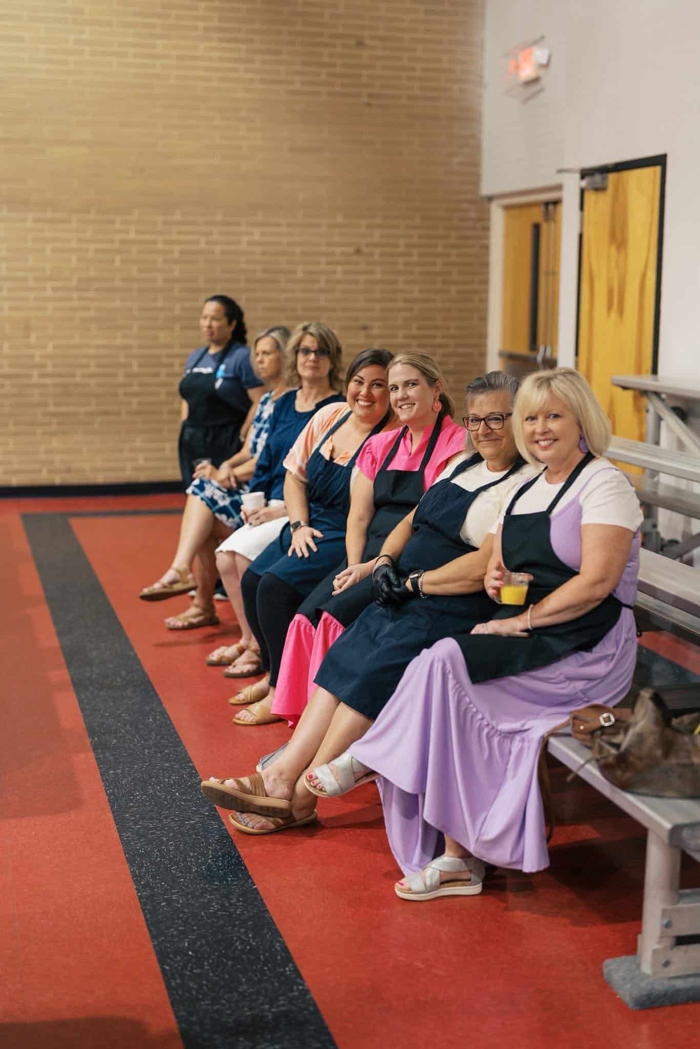 Six women seated on bleachers beside a red carpet in a gym, facing the camera.