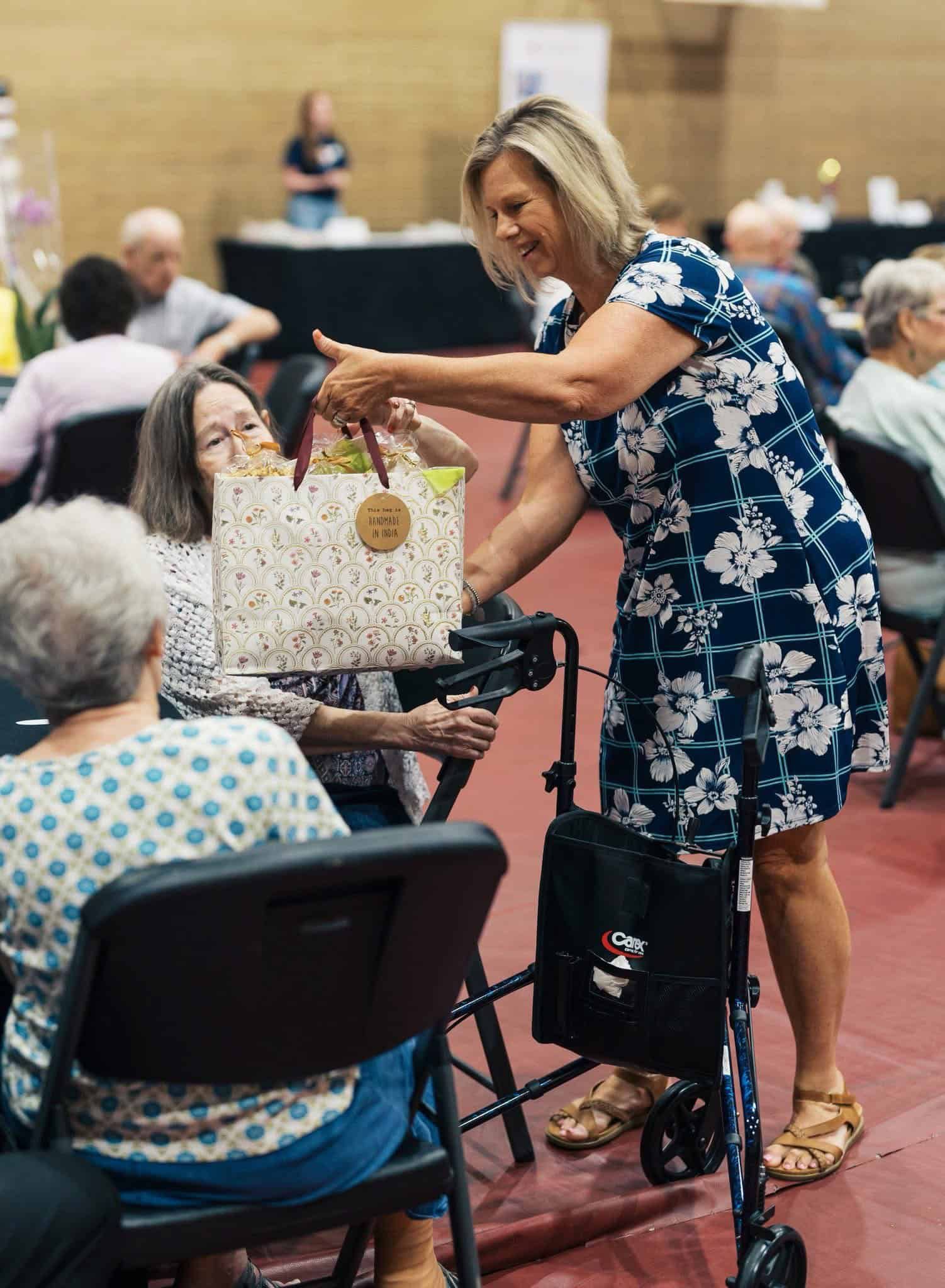 Woman in blue floral dress handing a gift bag to an seated person in a community hall