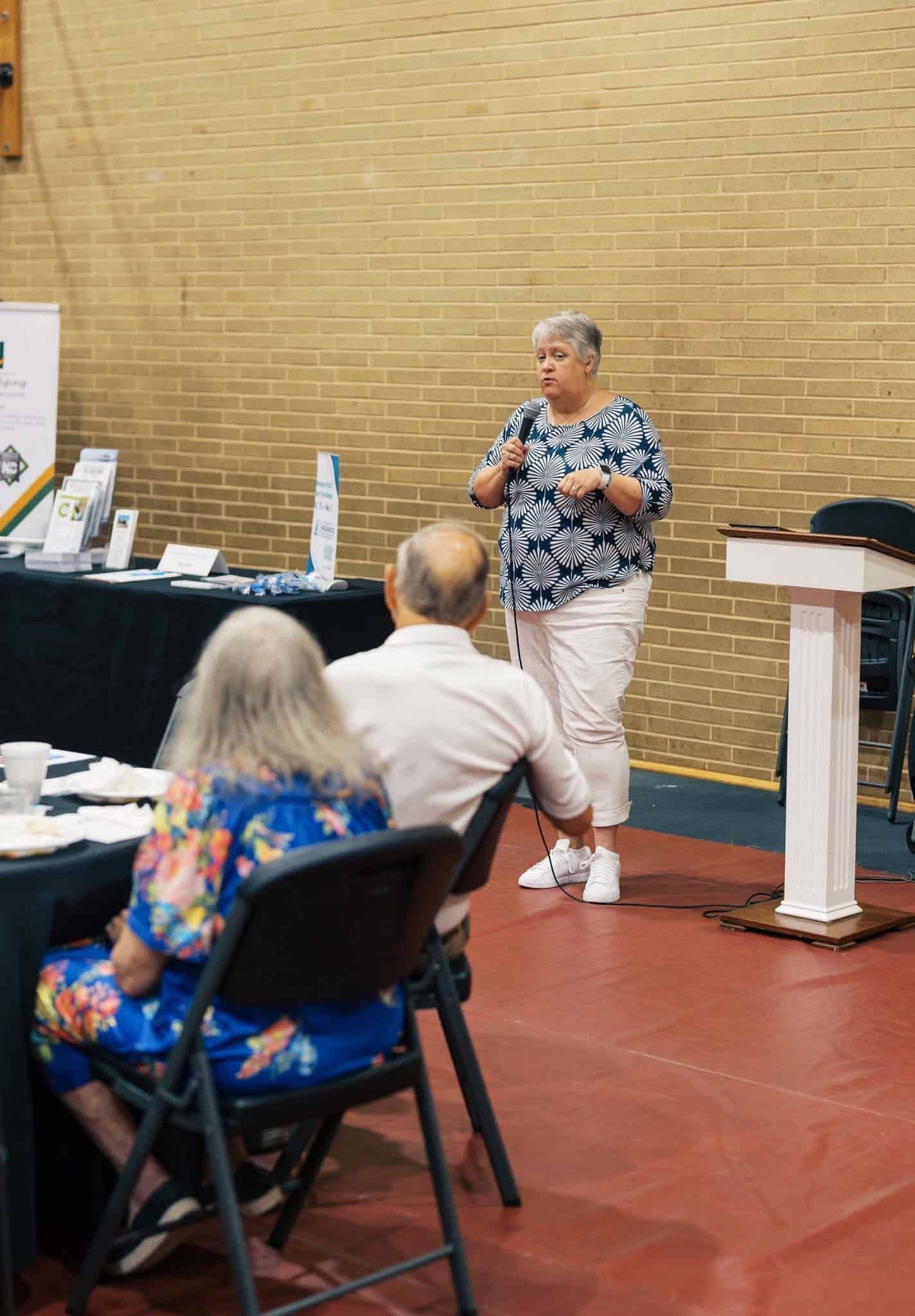 Speaker presenting in a meeting room beside a podium, with seated attendees in the foreground.