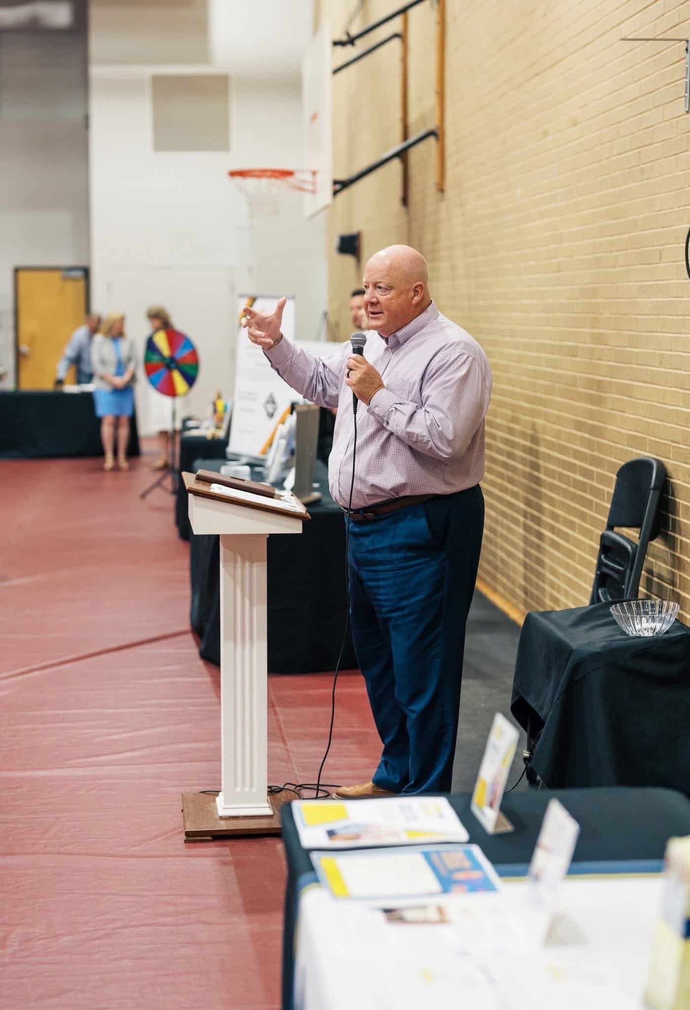 Speaker at a podium addressing an indoor gym audience, with papers and a microphone nearby.