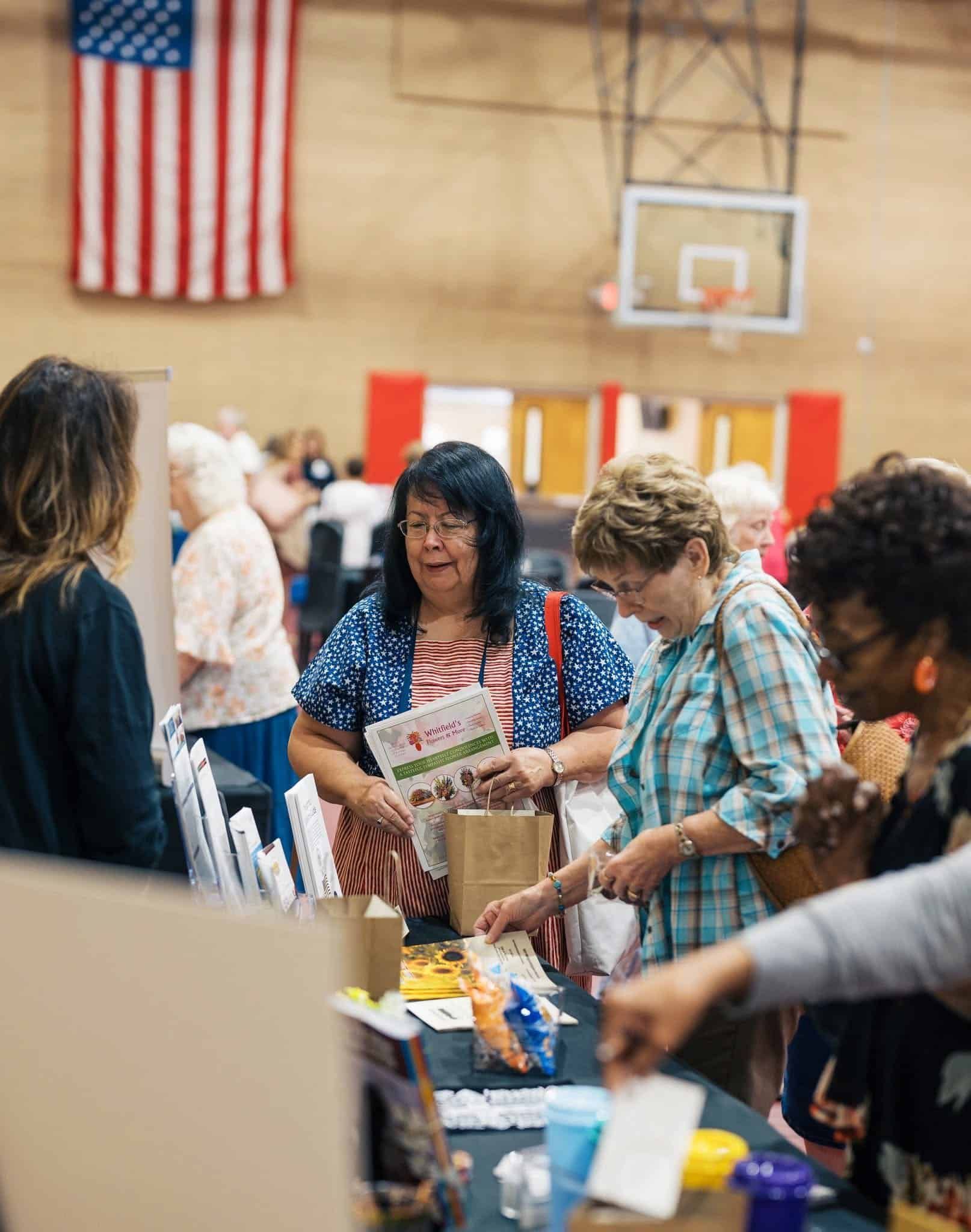 People browsing items at a table during a community indoor event with a basketball hoop and American flag in the background