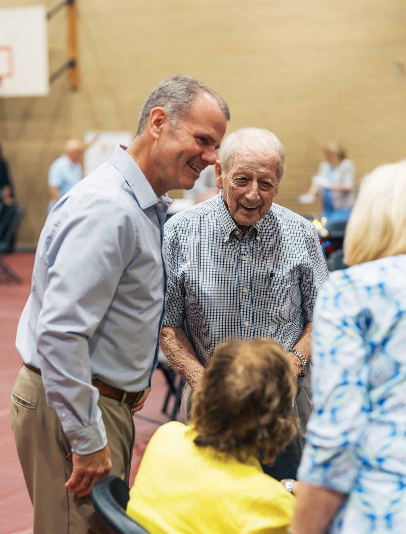 Man in blue shirt smiling and talking with an older man at an indoor gathering