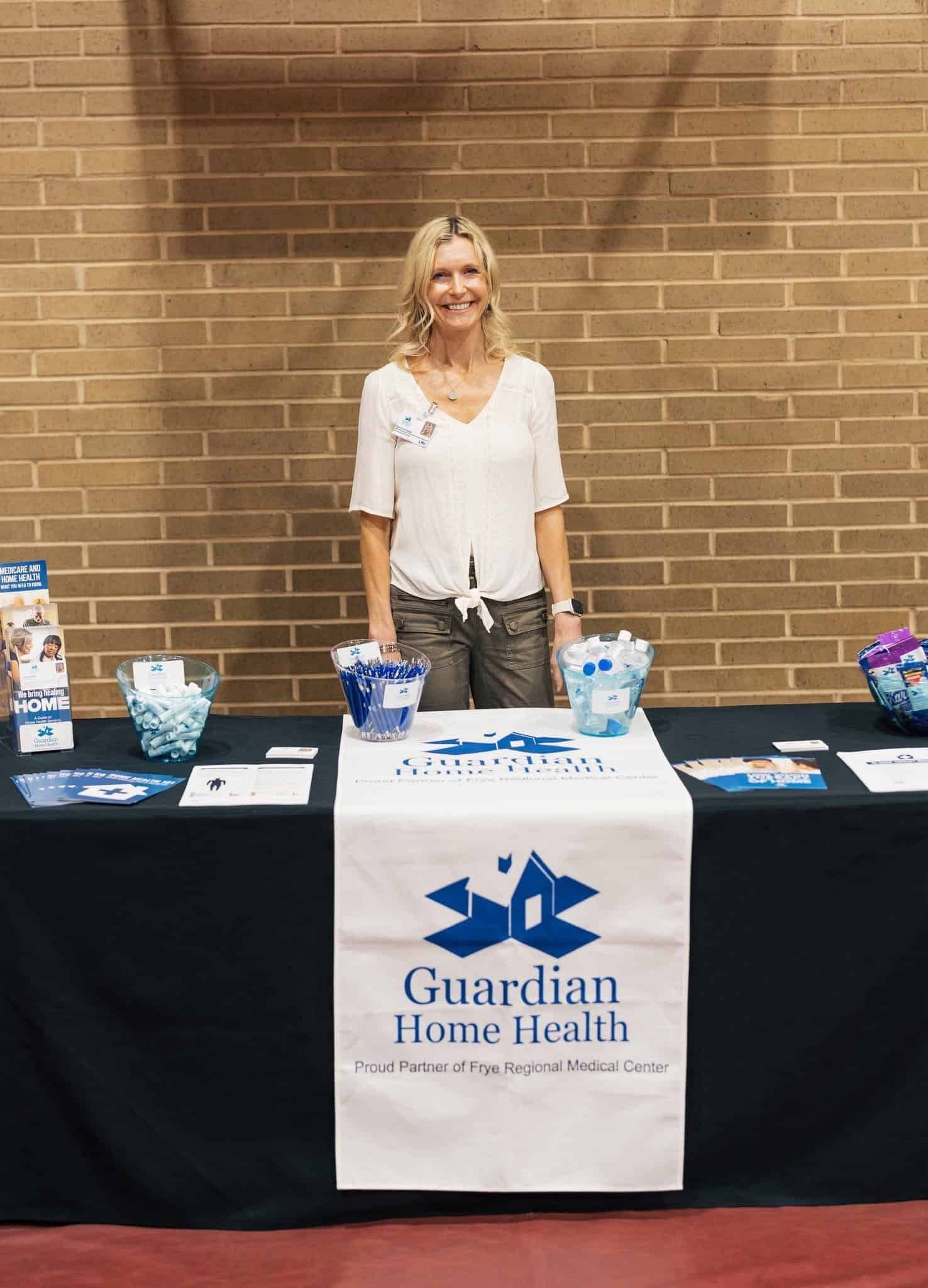 Woman standing behind Guardian Home Health table with brochures and promotional items against a brick wall