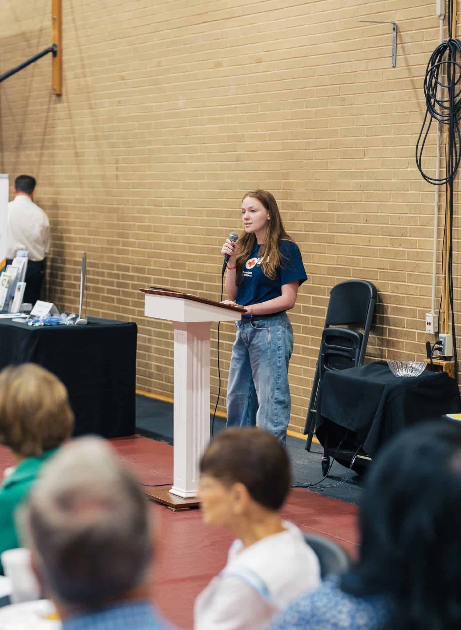 Woman speaking at a podium in a gymnasium, with audience seated in the foreground.