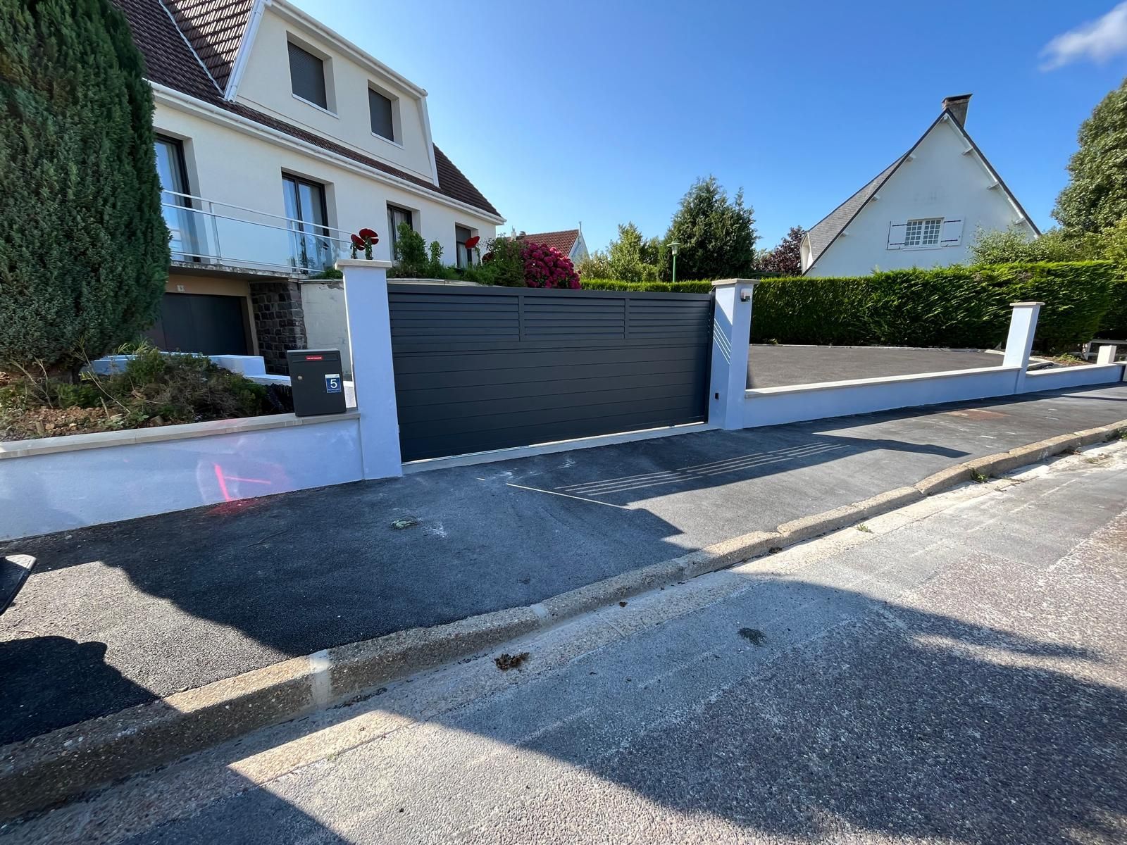 Un portail gris foncé devant une maison blanche avec une allée en asphalte noir par une journée ensoleillée.
