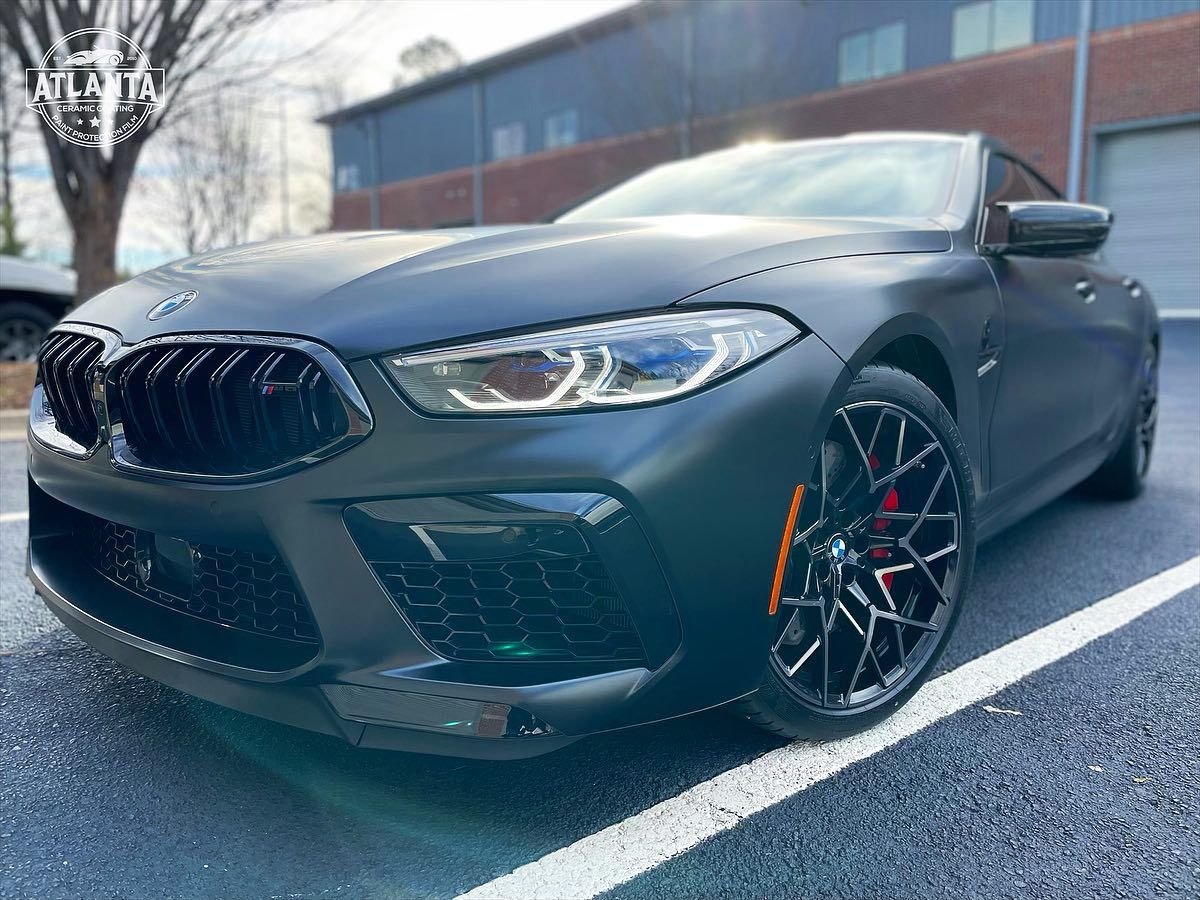 Matte black BMW M8 coupe parked in a parking lot. Red brake calipers visible.