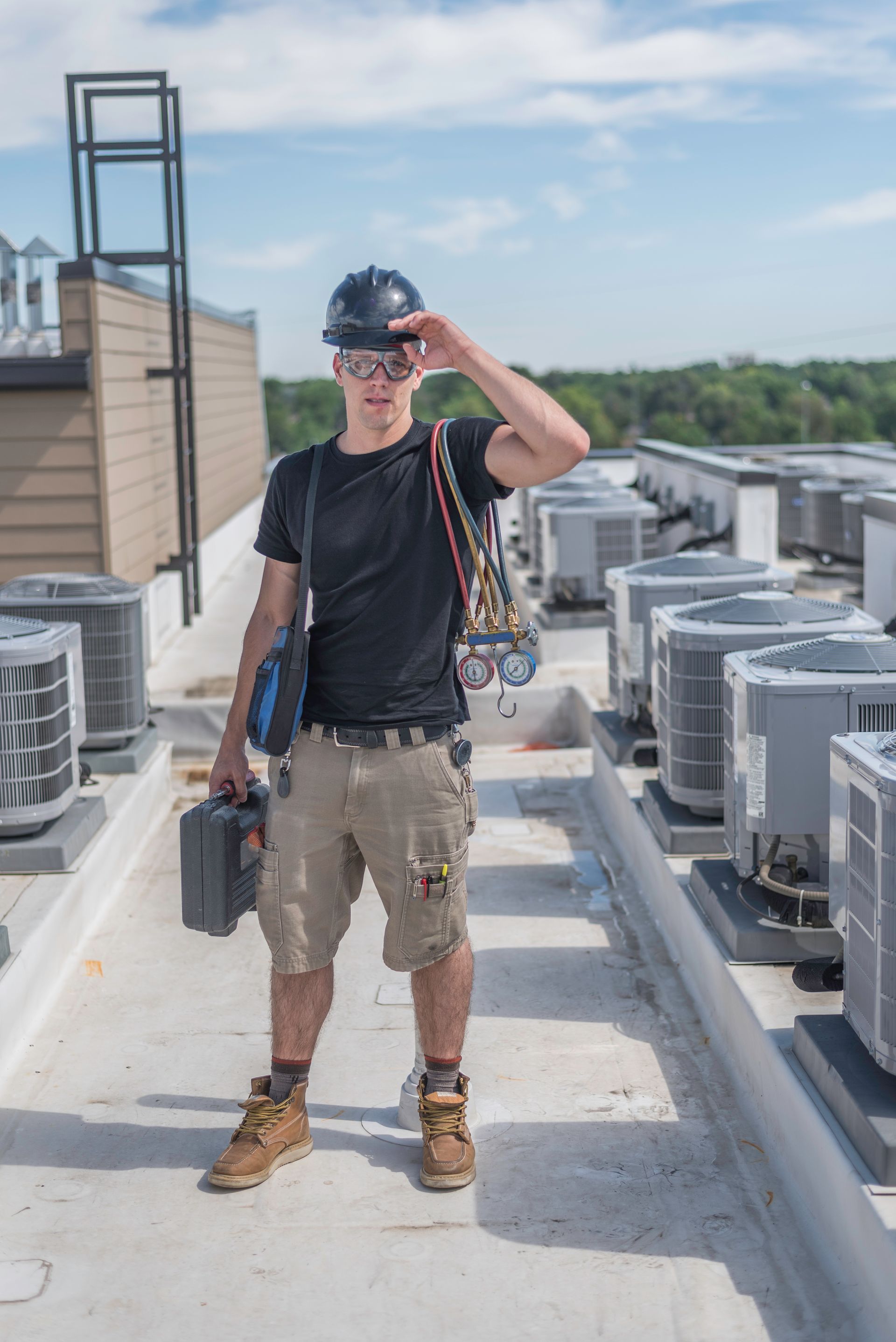 Construction worker on a rooftop with equipment, wearing a hard hat and sunglasses, saluting in sunny conditions.