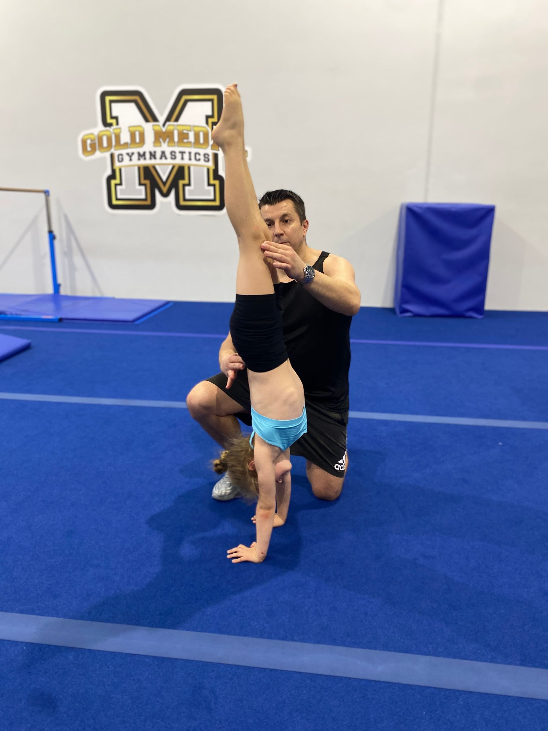A Man is Helping a Little Girl Do a Handstand on a Blue Mat — Gold Medal Gymnastics In Molendinar, QLD