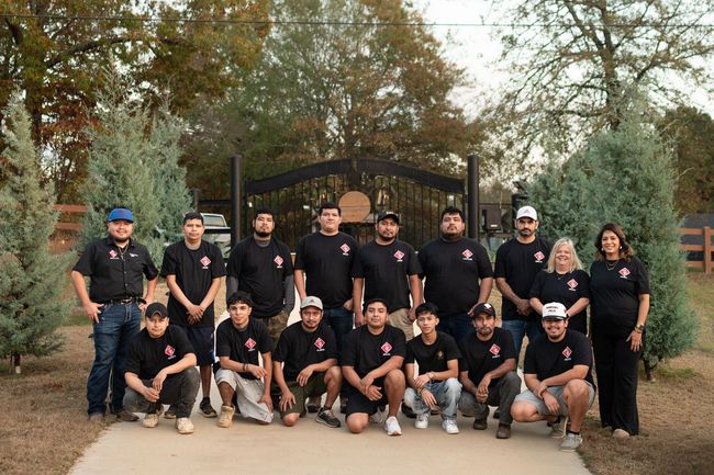 Group of people in black shirts posing outdoors in front of a gate and trees.
