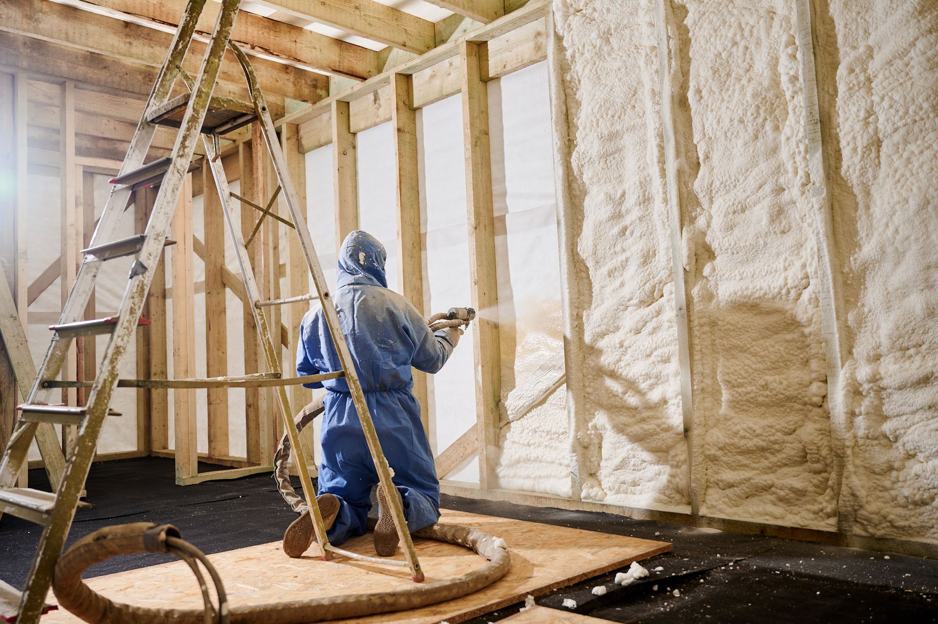 Worker in blue coveralls spray-foaming insulation inside an unfinished attic with exposed wooden beams.