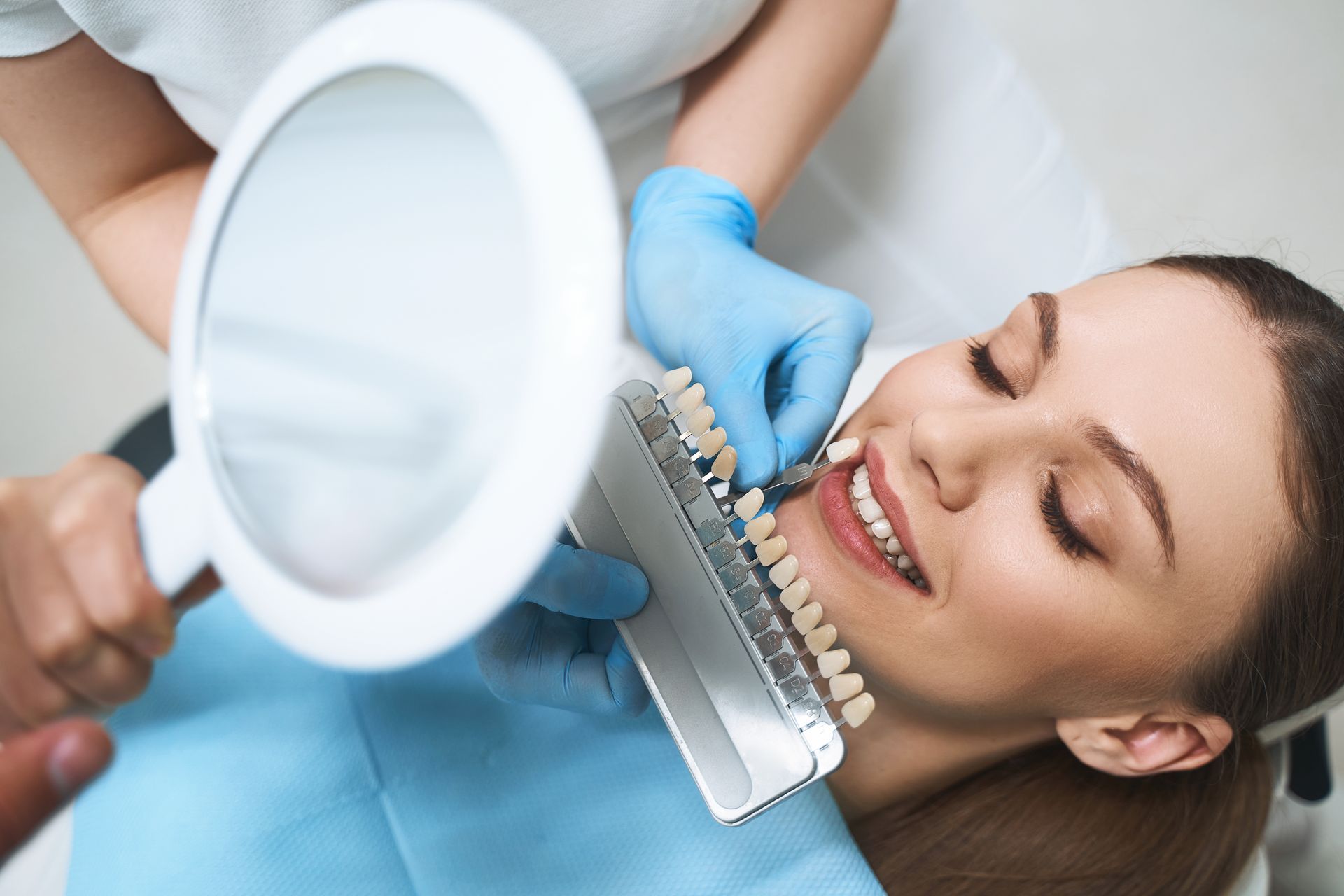 Dentist comparing teeth shade to a guide using a magnifying glass, patient smiling in dental chair.