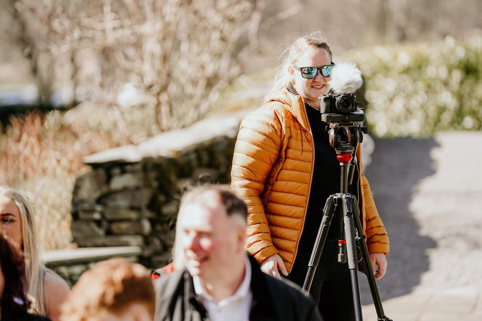 Sophie in an orange jacket stands by a tripod camera outdoors at Lake District Wedding, wearing sunglasses.