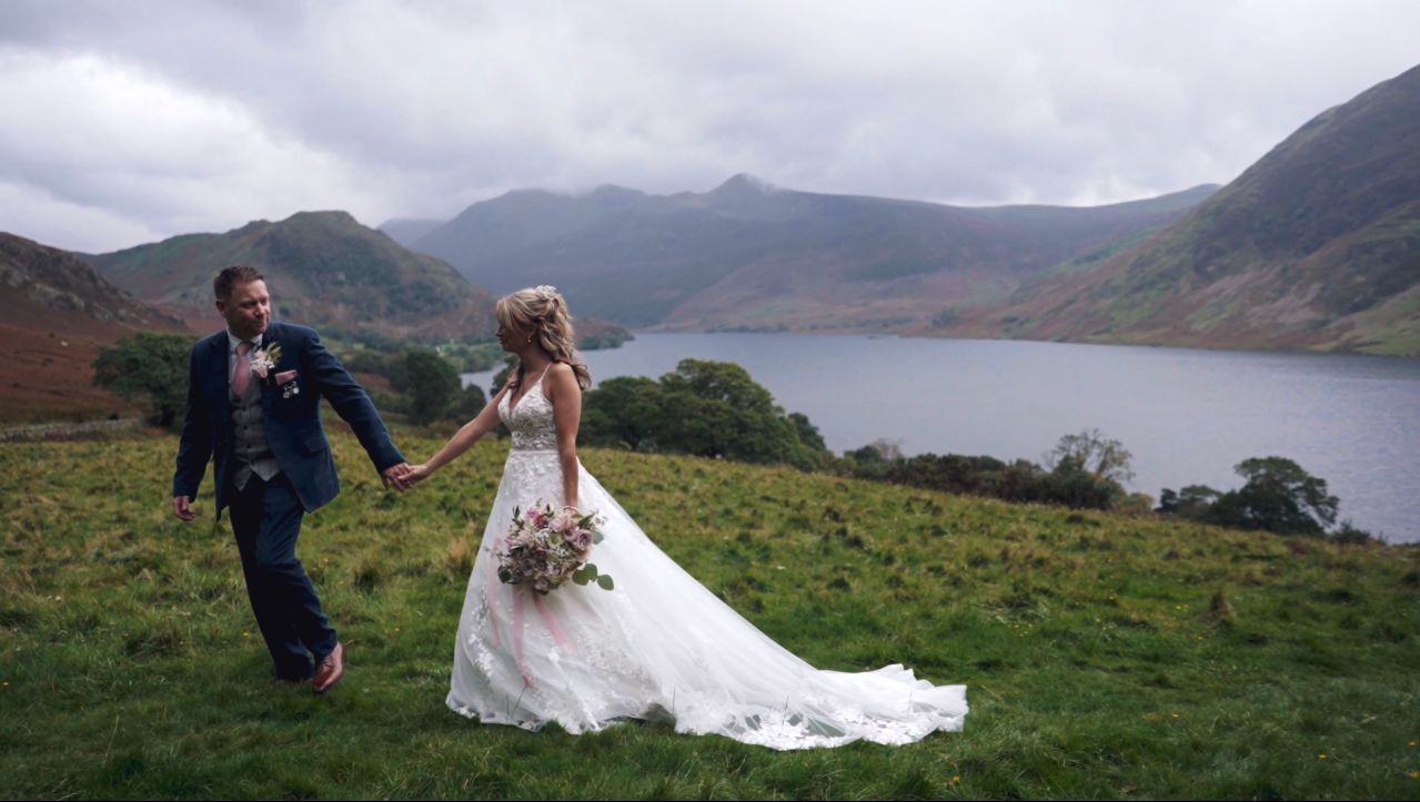 Bride and groom holding hands on a grassy hillside overlooking  Lake Windermere  and mountains