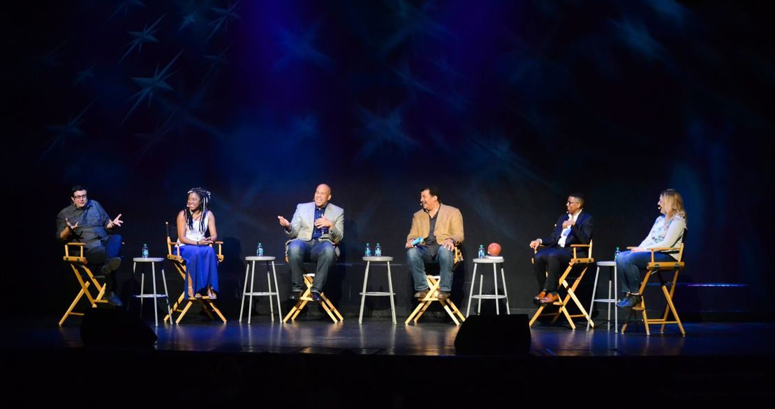 Six people seated on stools in a dim blue-lit stage panel discussion