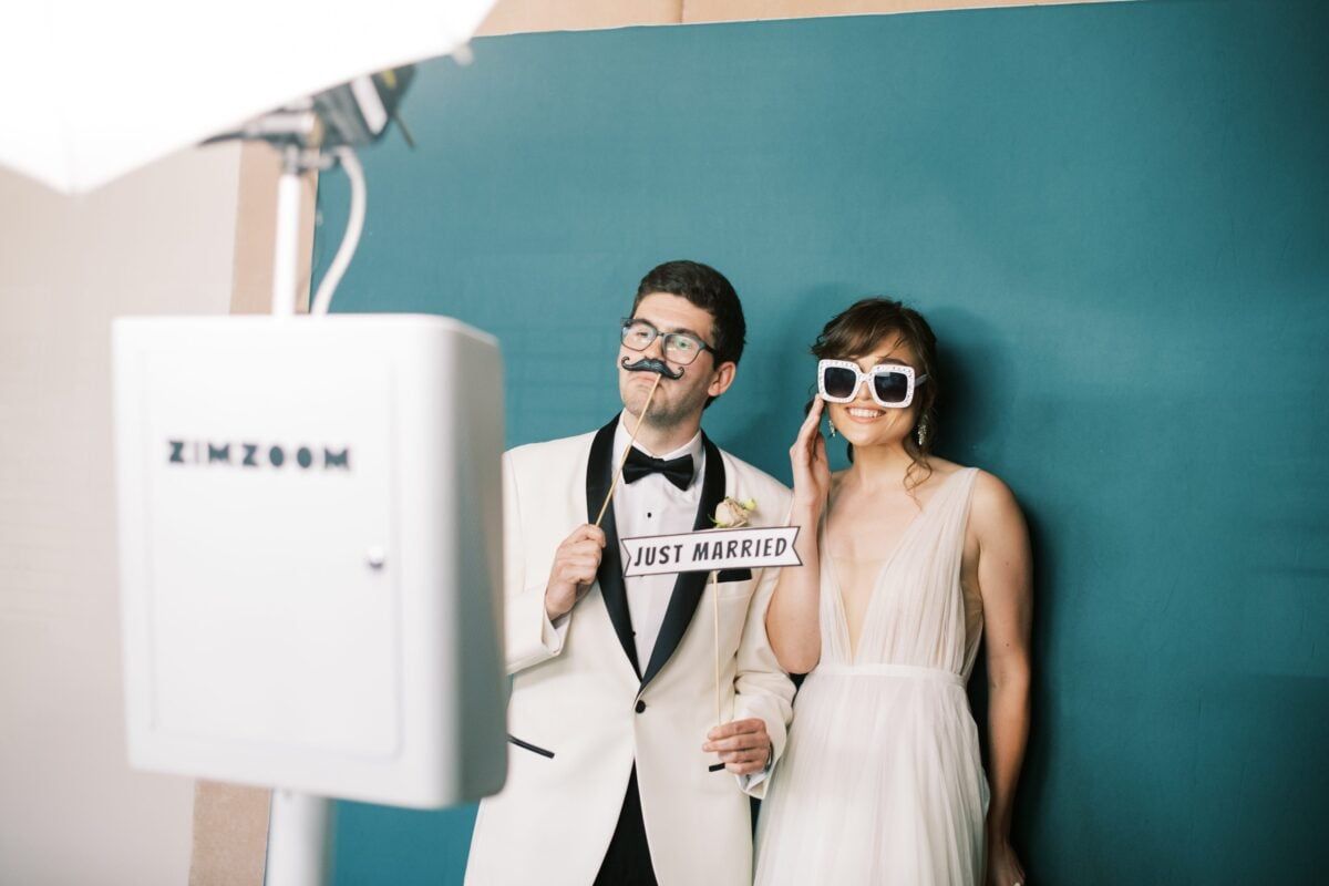Two people in sunglasses pose beside a white photo booth against a teal wall.