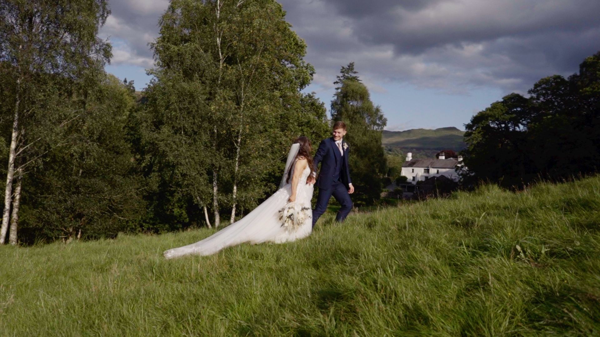 Bride and groom walking near Coniston, through a sunlit grassy field with trees and a house in the background.