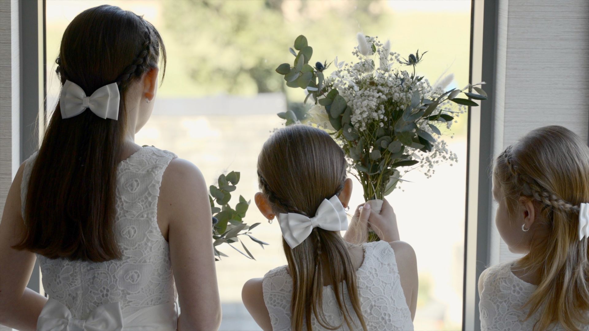 Three children in white dresses with bows look out a window at a vase of flowers.