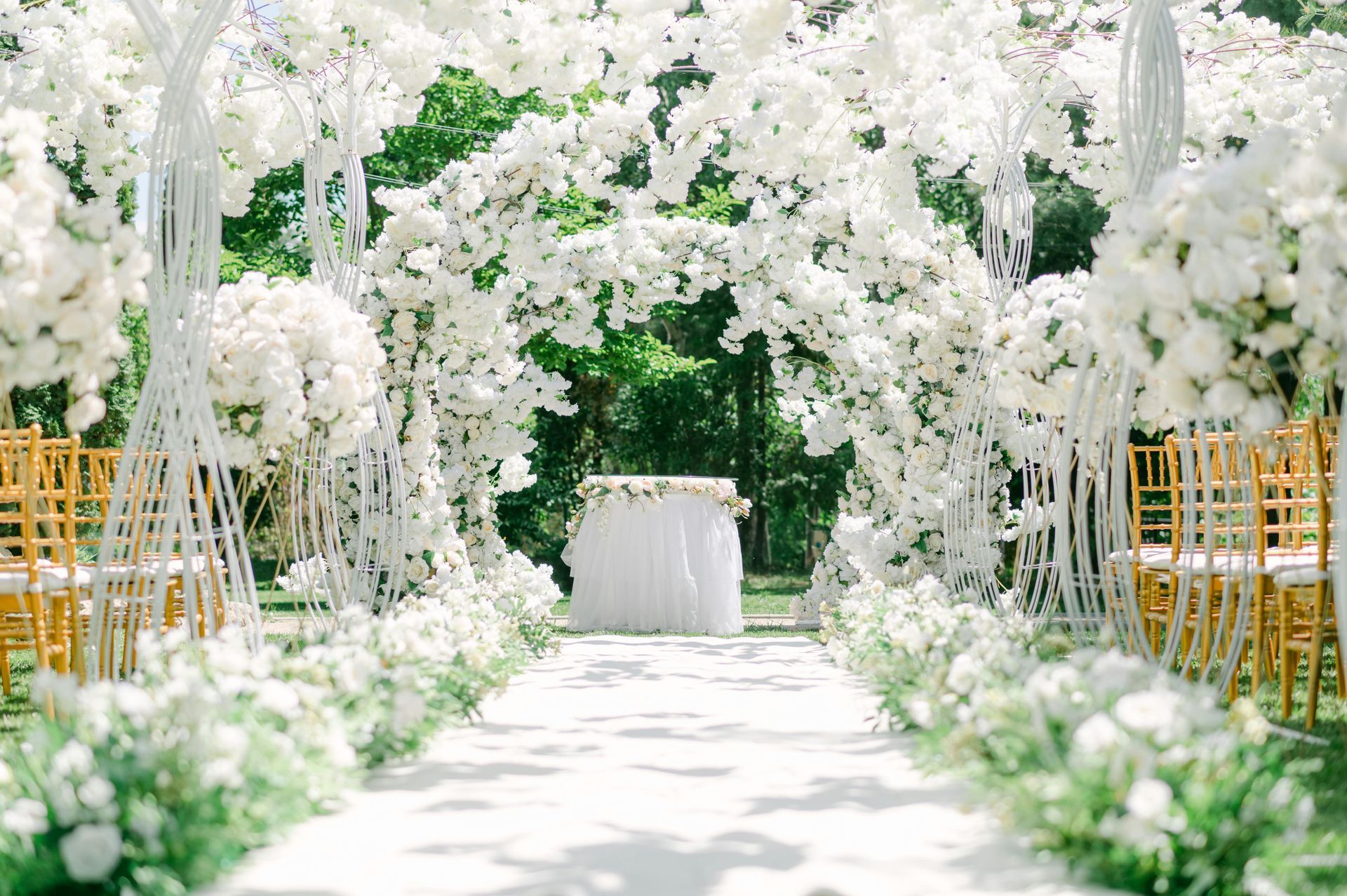 White floral wedding aisle with lush archway, greenery, and gold chairs leading to a small table at the altar