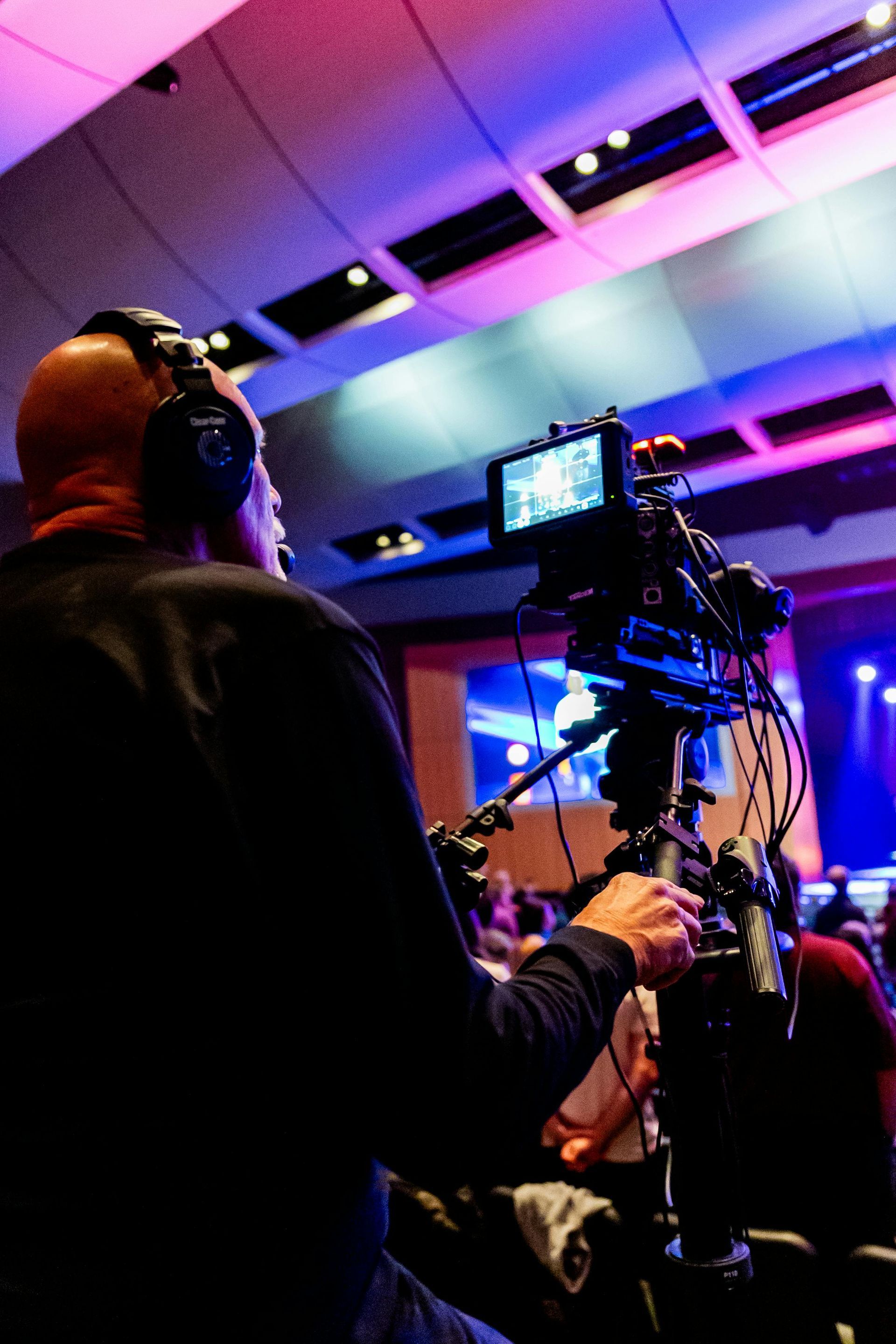 Camera operator filming a concert, with purple stage lights and a monitor on a tripod