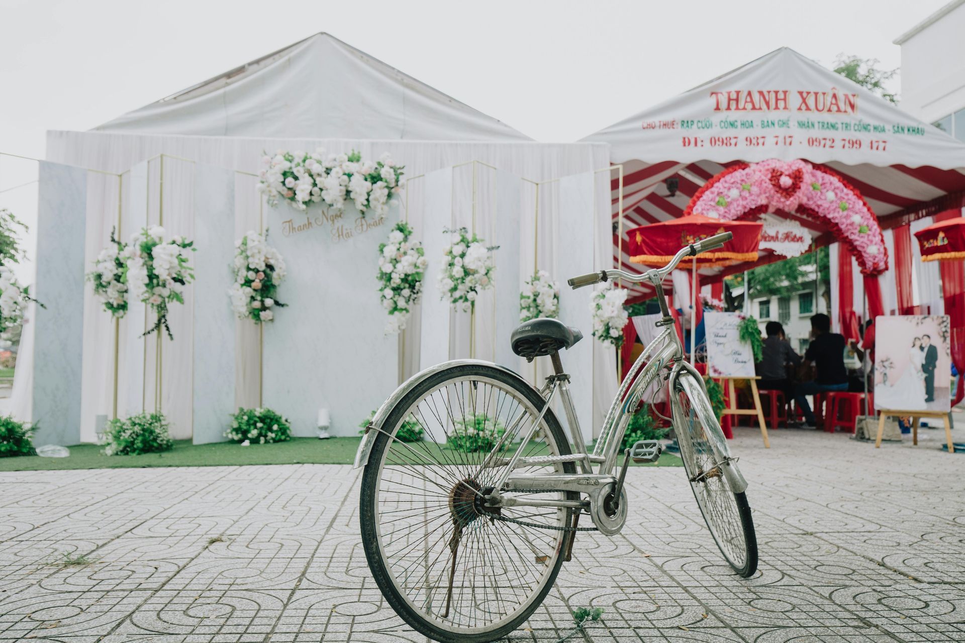 White event tent with floral decorations and a decorative bicycle in front, next to a red wedding-style canopy.