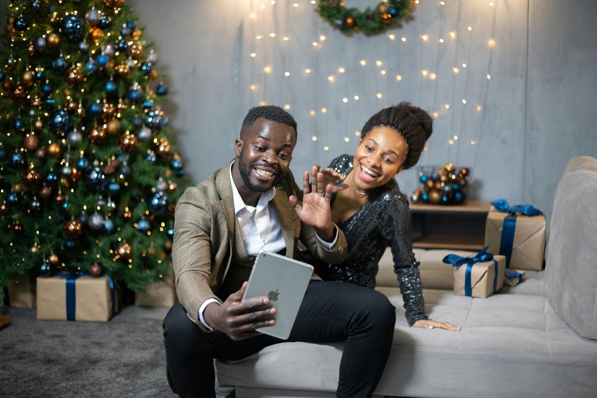 Couple waving on a couch beside a decorated Christmas tree and warm holiday lights
