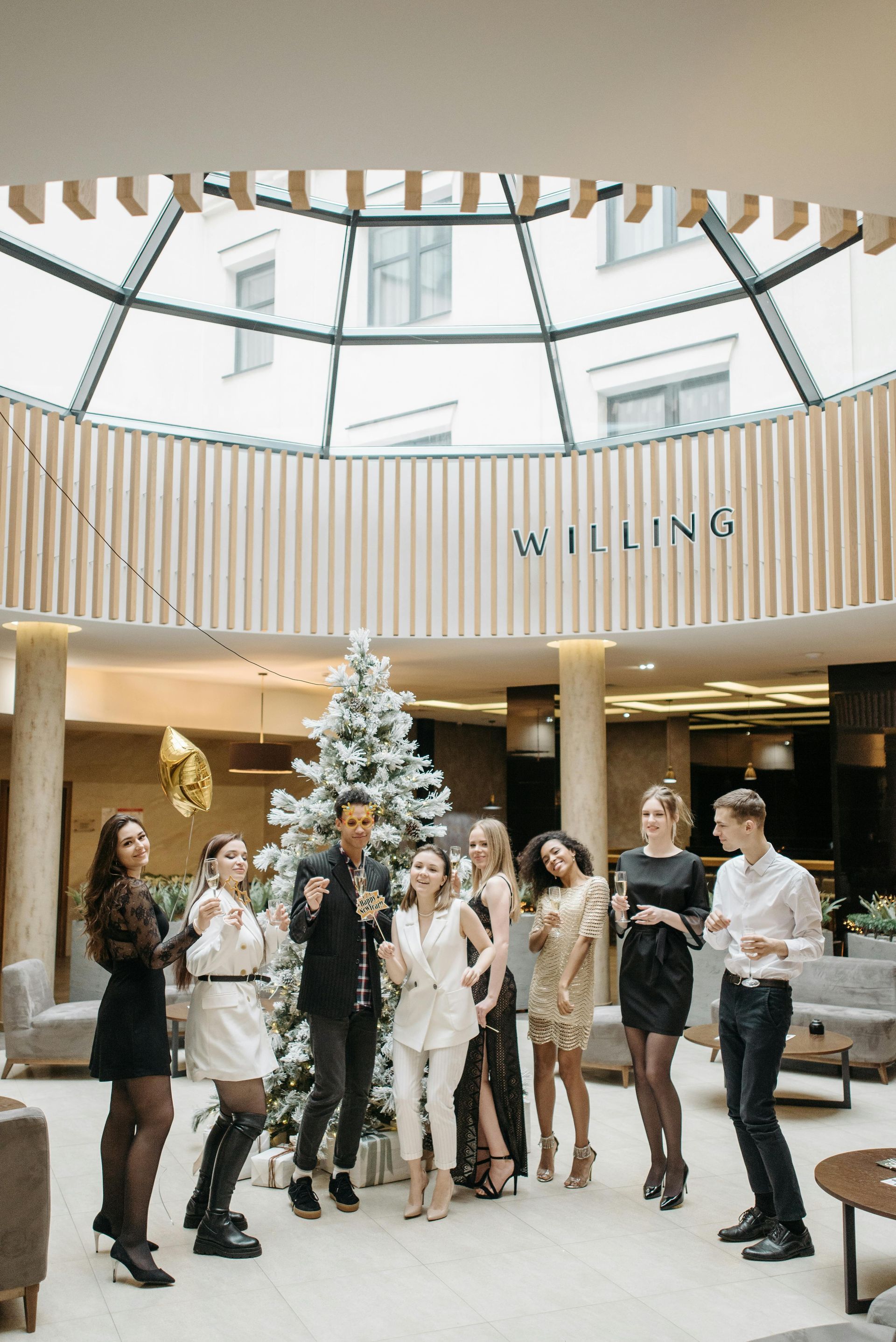 Group of people gathered around a decorated Christmas tree in a bright indoor atrium.