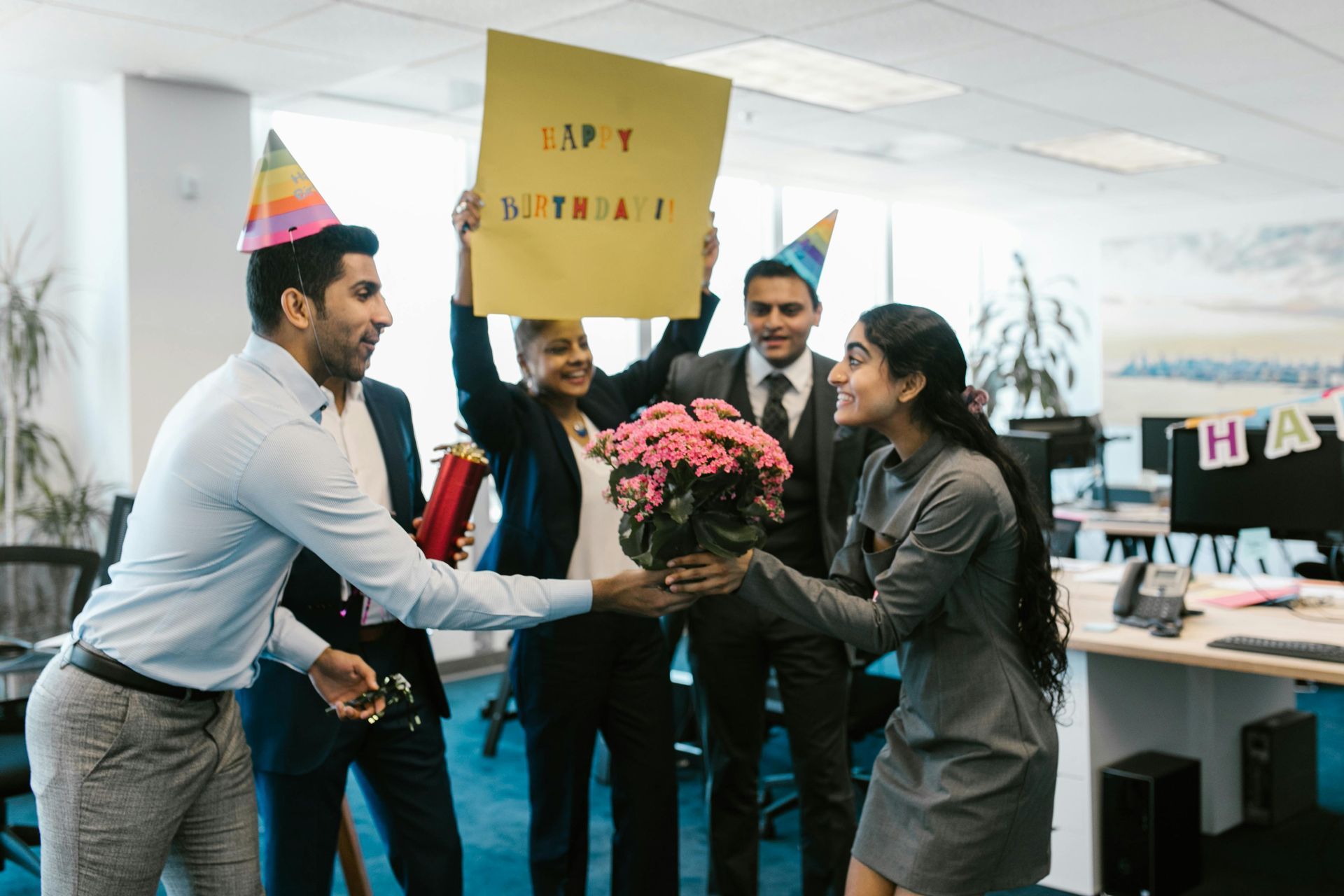 Coworkers in party hats celebrate in an office, holding flowers and a sign.
