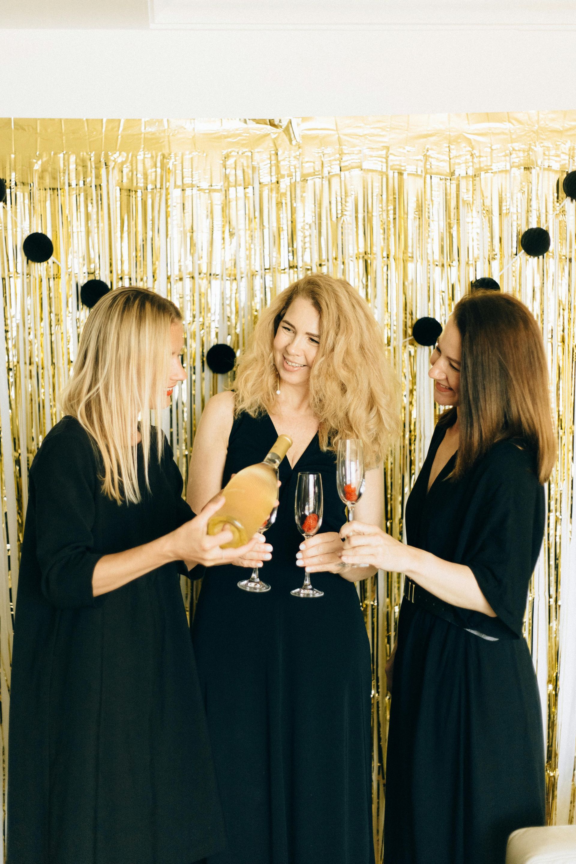 Three women in black dresses holding champagne glasses at a gold tinsel party backdrop