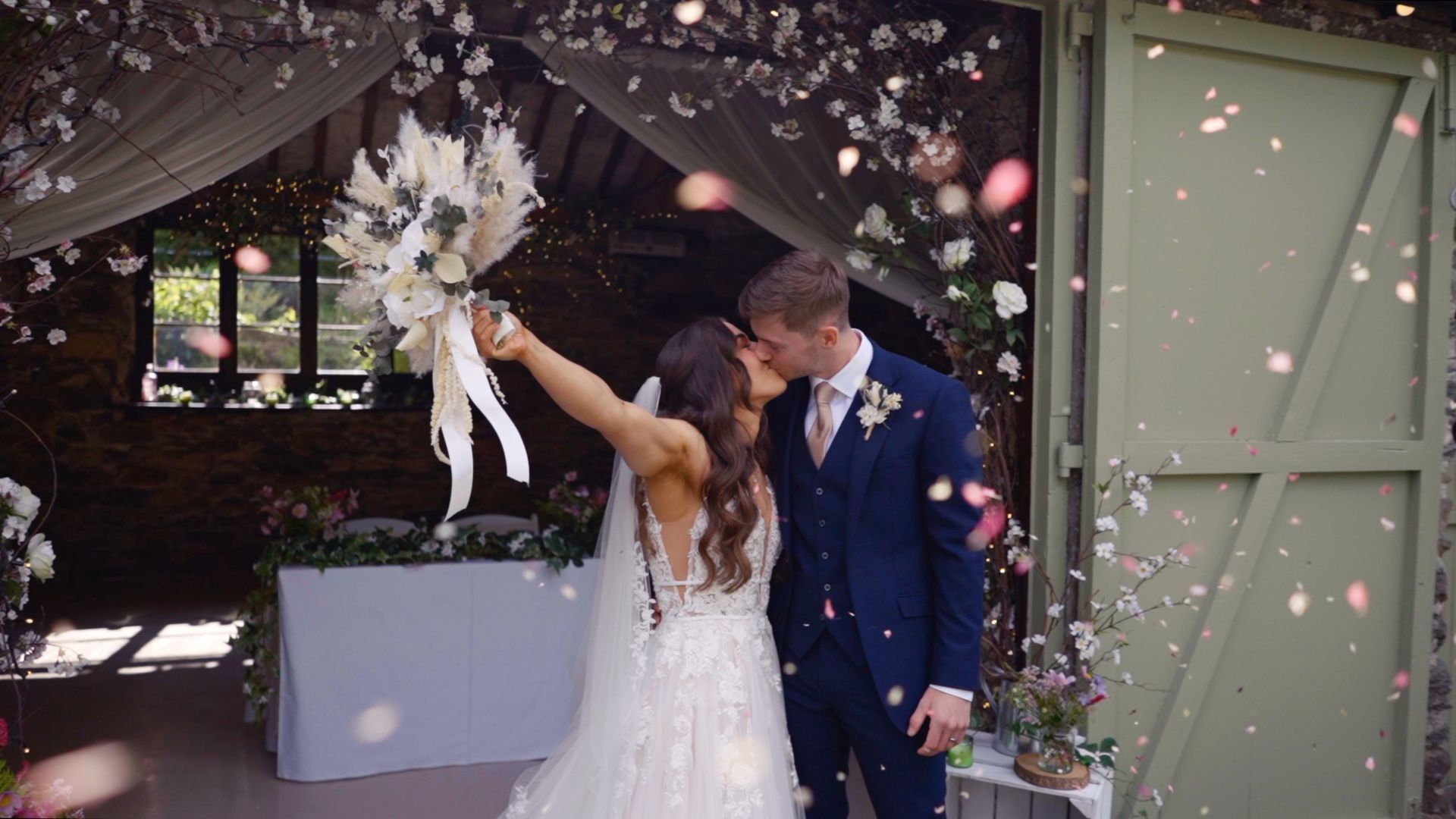 Bride and groom kissing under floral arch with falling petals in Windemere