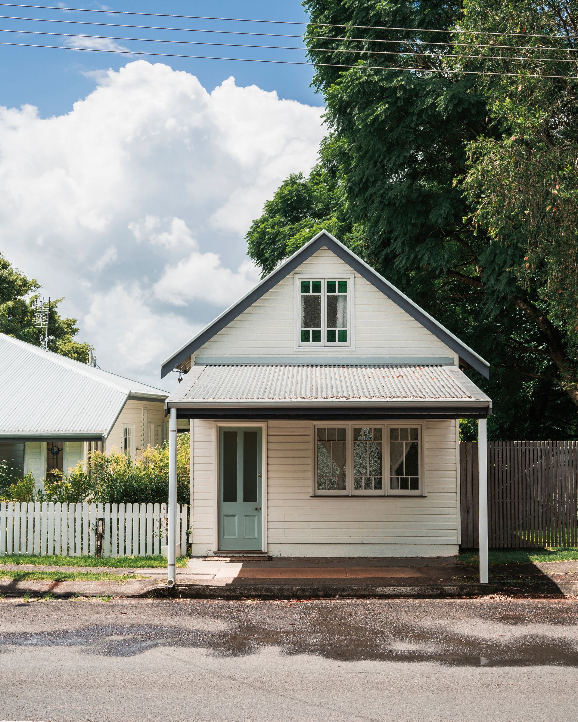 Small white cottage with green-trimmed gable, front porch, and large tree beside a street
