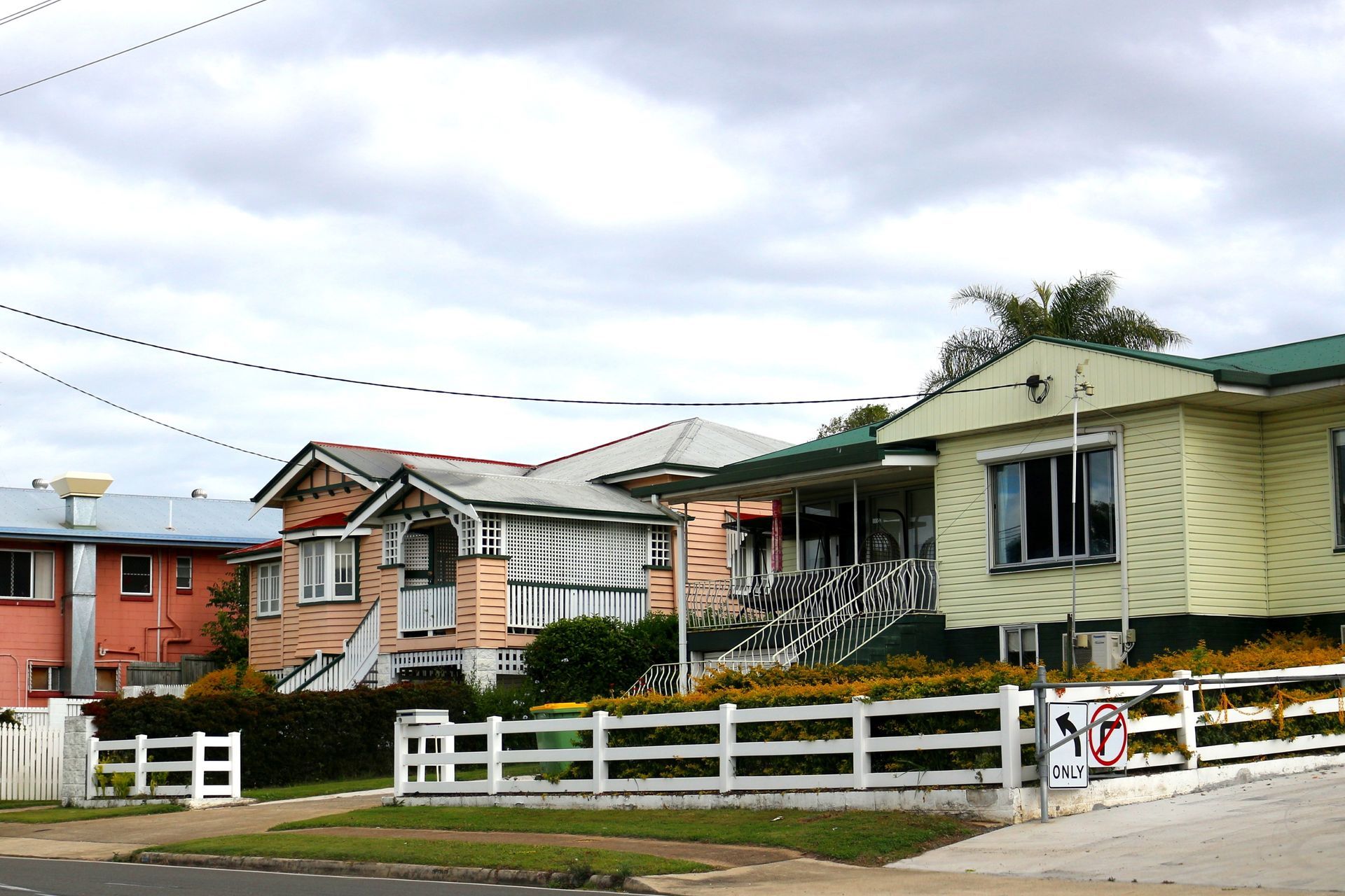 Row of damaged houses with white fences along a street under a cloudy sky