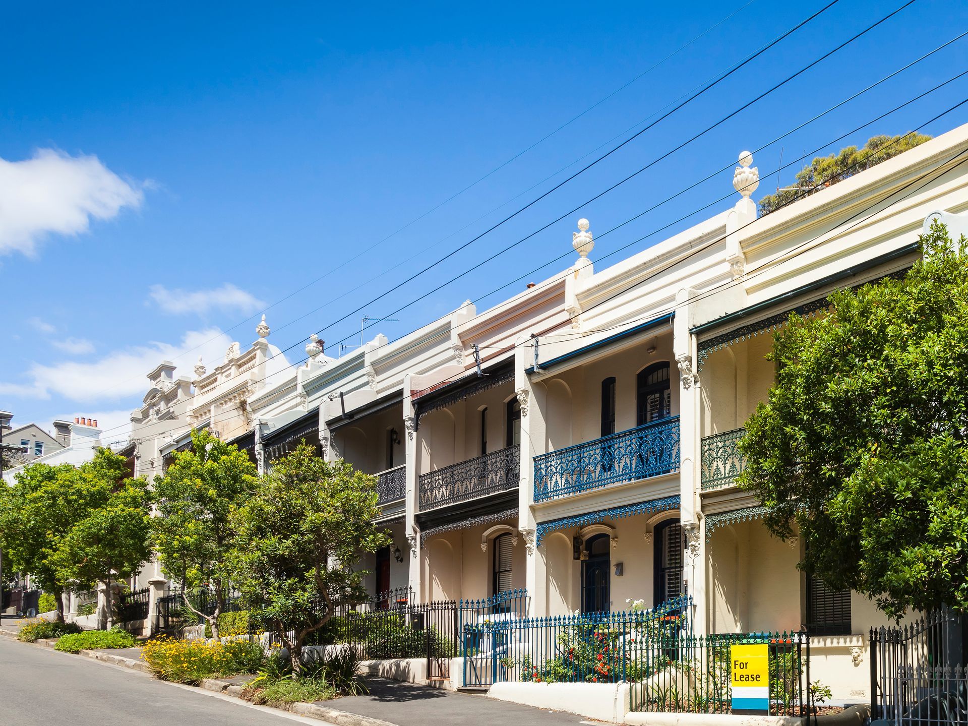 Row of cream-colored townhouses with blue balconies along a sunny tree-lined street