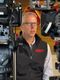 Man speaking beside video cameras in a studio, wearing glasses and a black vest with a red logo.