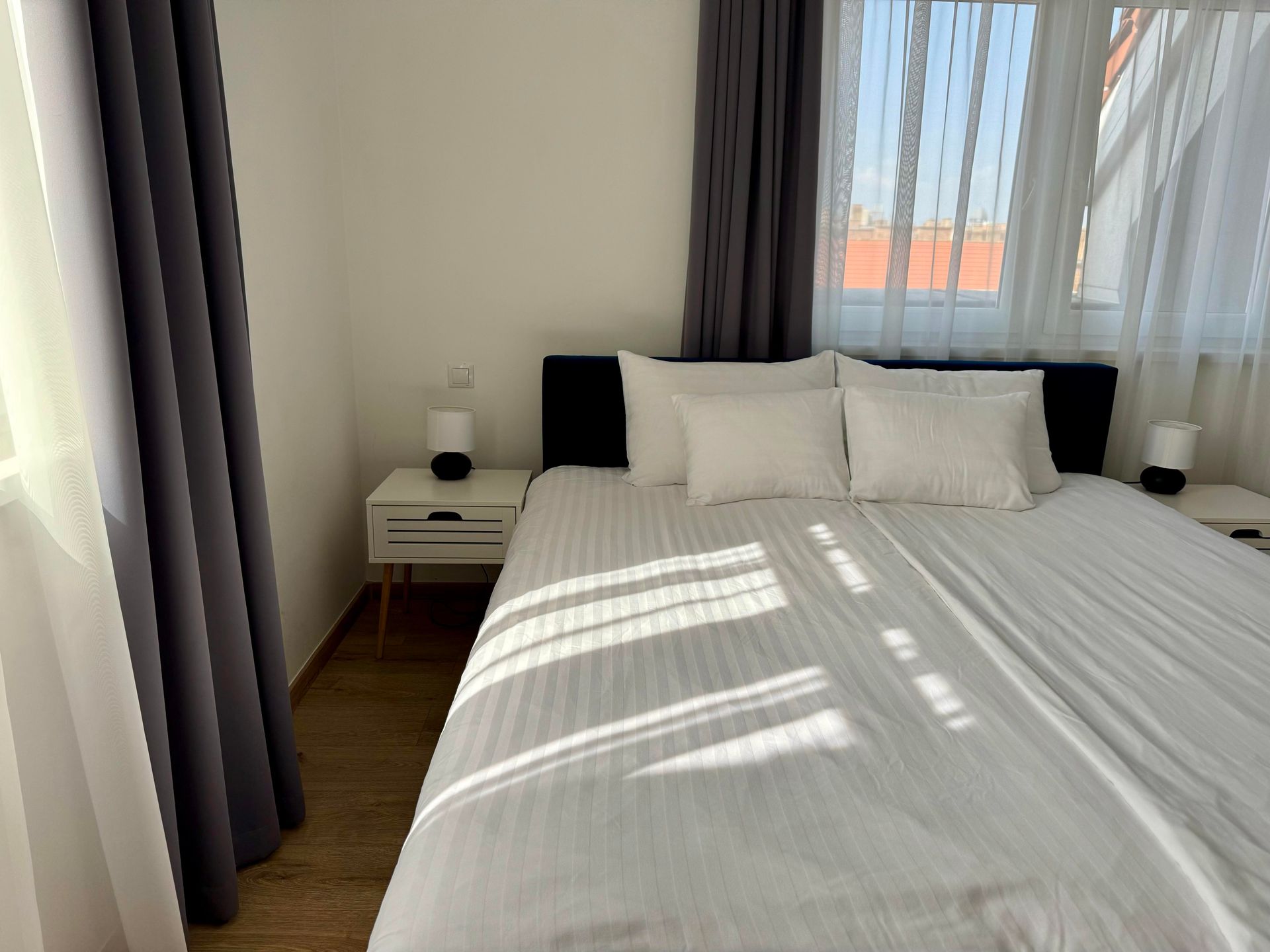 Bedroom with a bed, nightstands, curtains, and windows. Sunlight streams in.