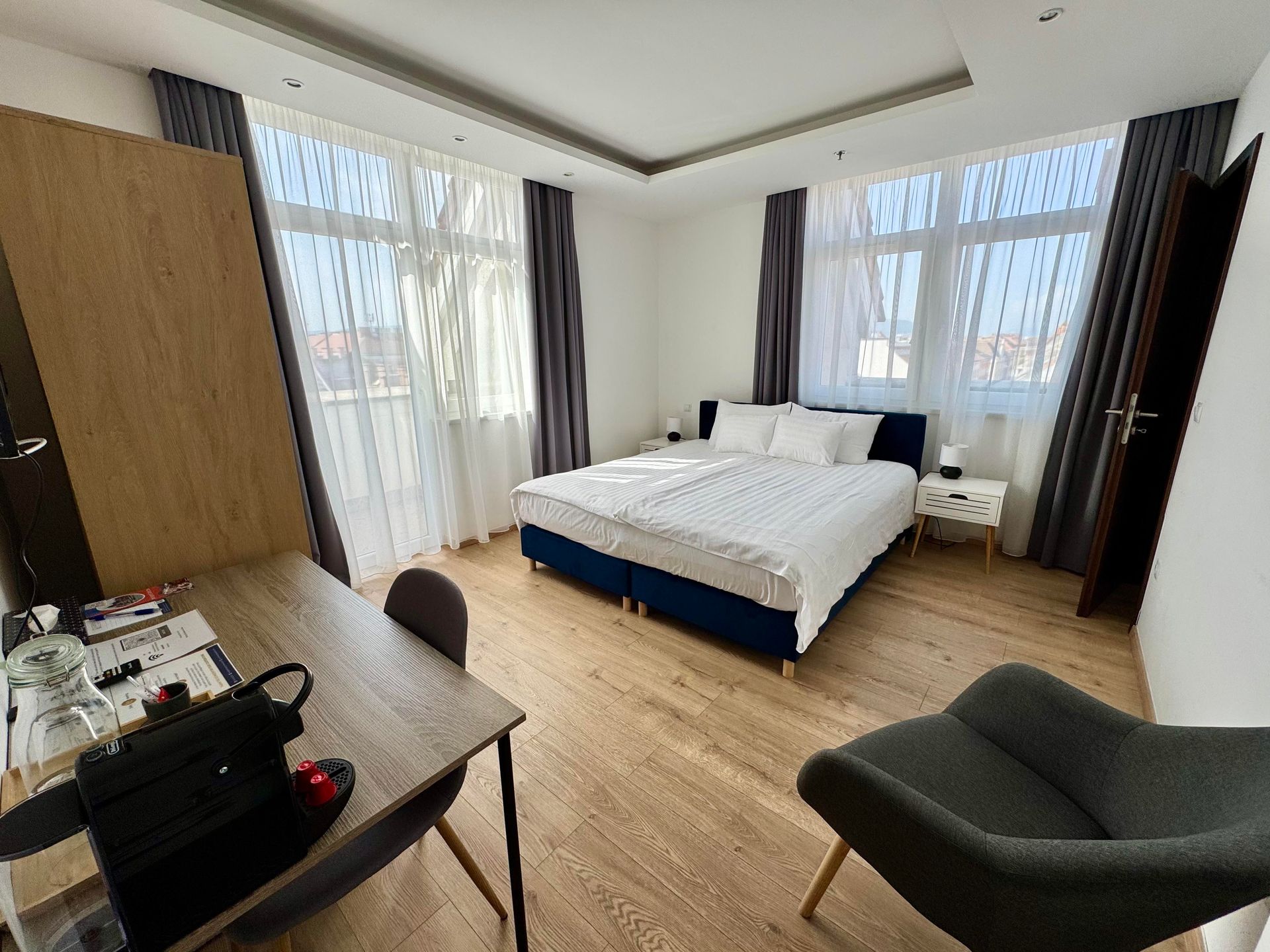 Bedroom with large bed, desk, and armchair. Windows with curtains and bright natural light.