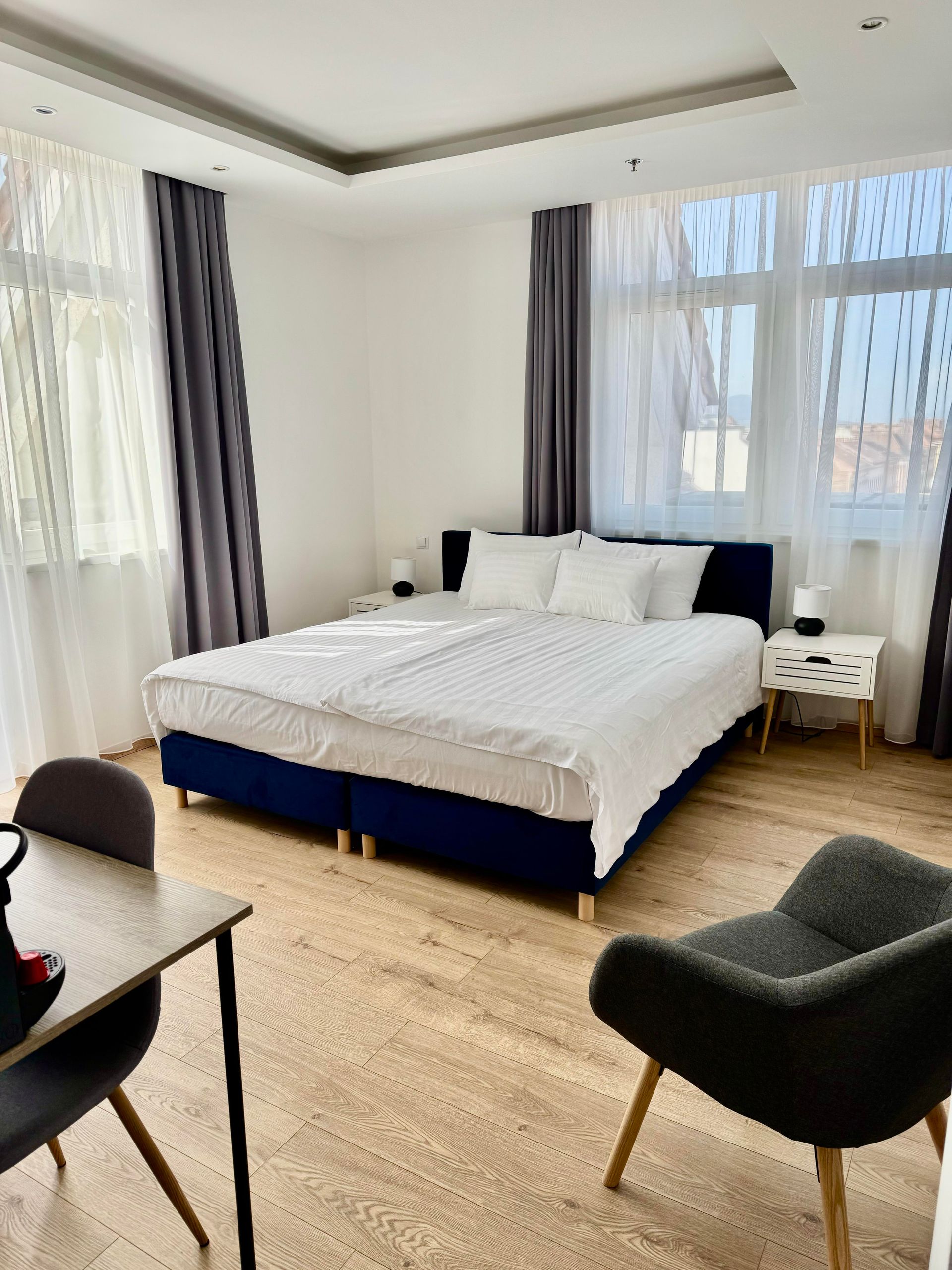 Bedroom with a large bed, two nightstands, a desk, and a chair. Gray curtains frame the windows.